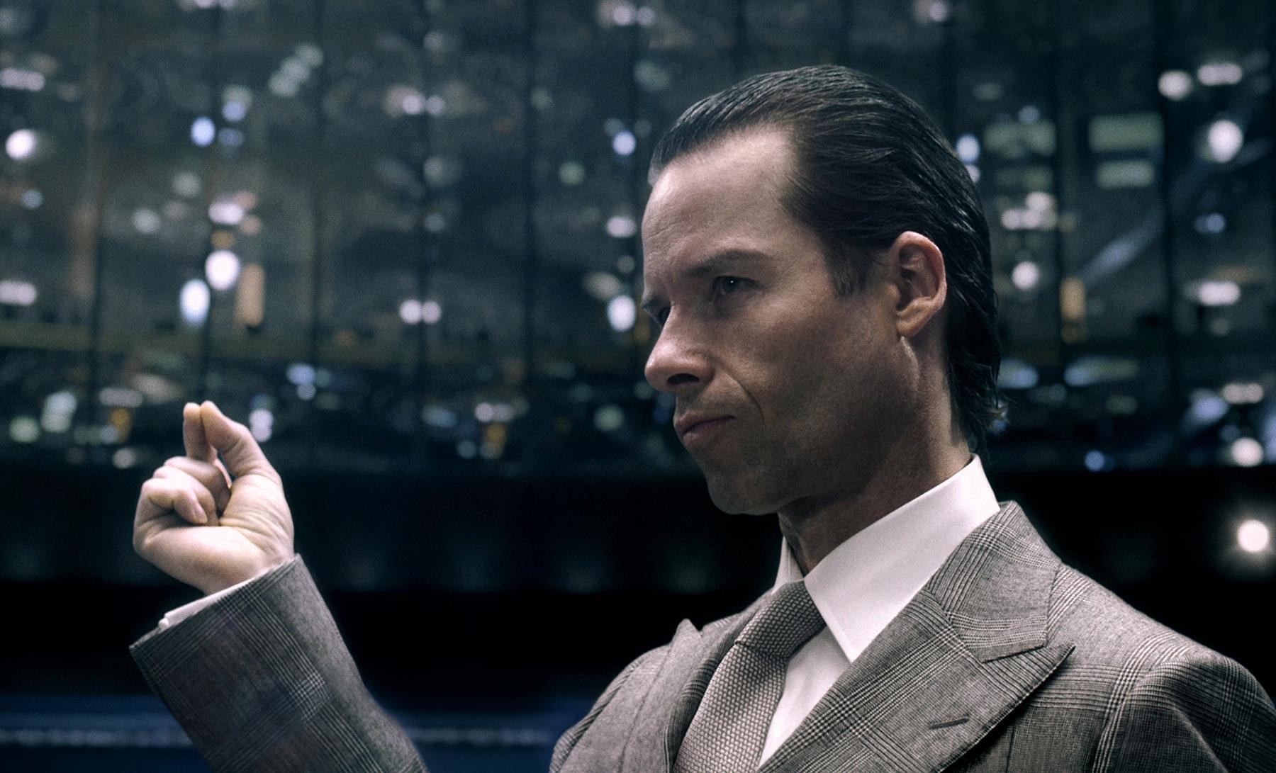 A close-up shot of Peter Weyland (Guy Pearce) in a tan suit and tie pinching his forefinger and thumb together while surrounded by an auditorium of people in a fake TED talk produced for the 2012 film Prometheus.