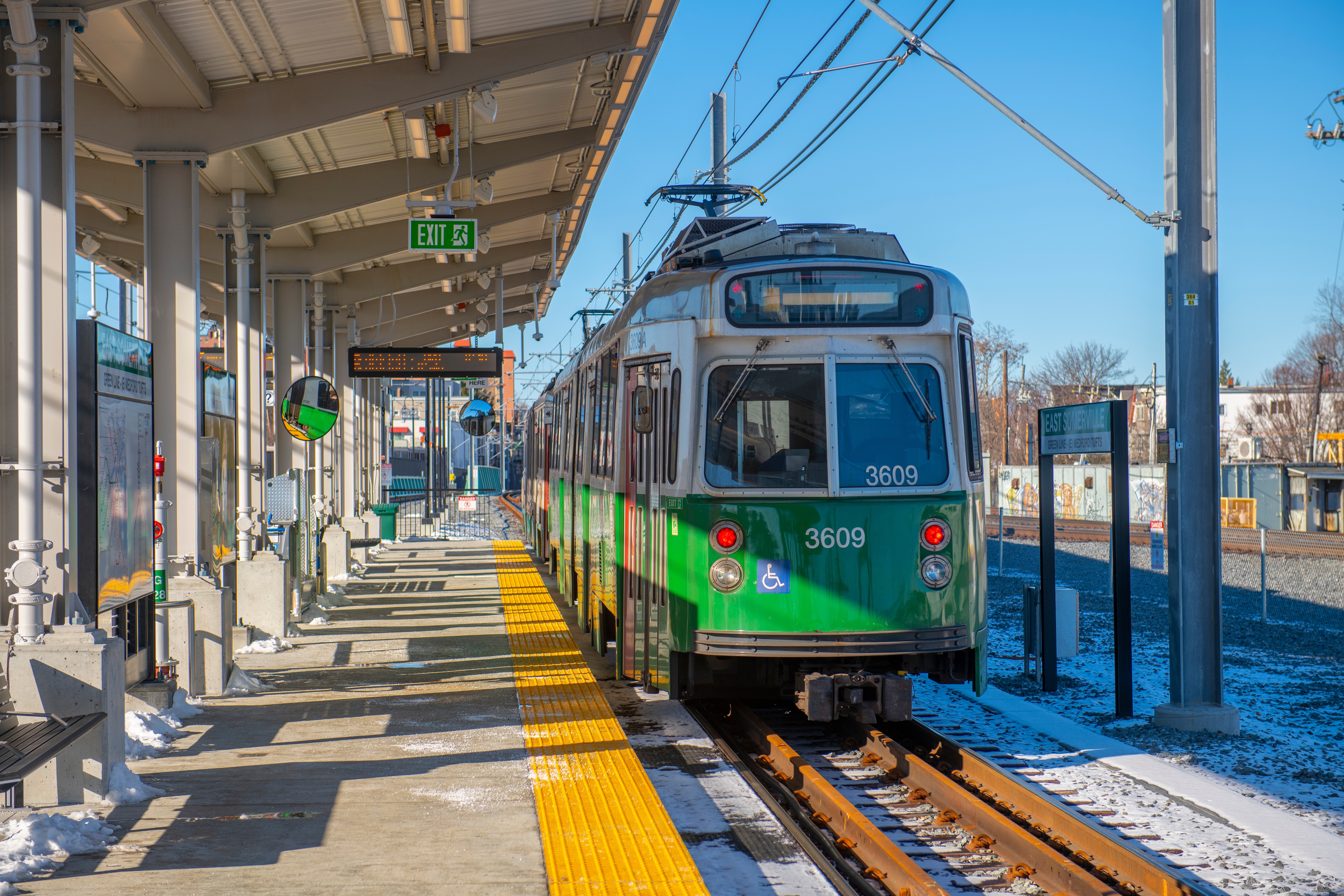 A green-hued above-ground subway train stops at an open-air station in the winter.