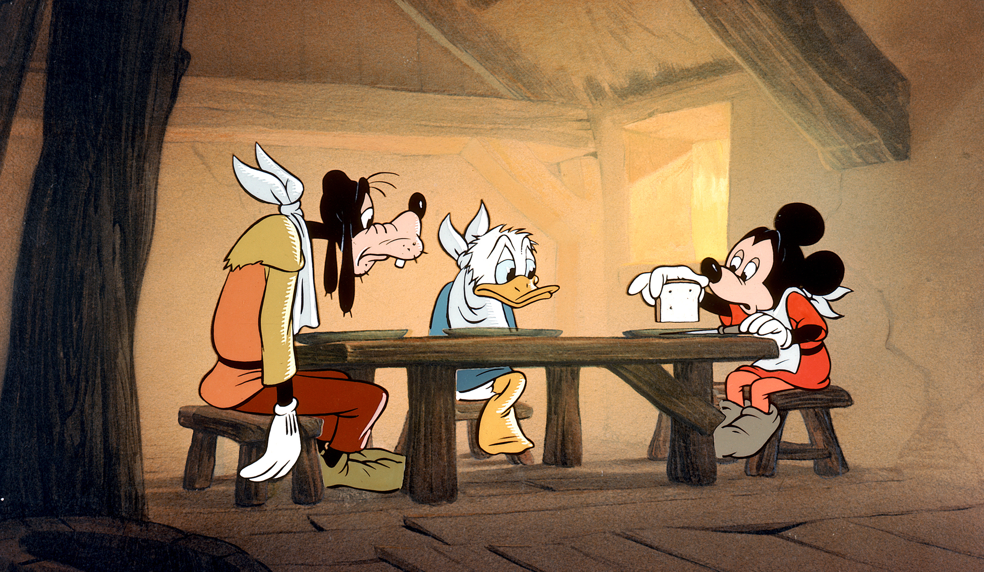 In a scene from the 1947 short Mickey and the Beanstalk, Goofy, Donald Duck, and Mickey Mouse slump hungrily over a wooden table in a bare room, wearing napkins around their necks and getting ready to split a single bean three ways