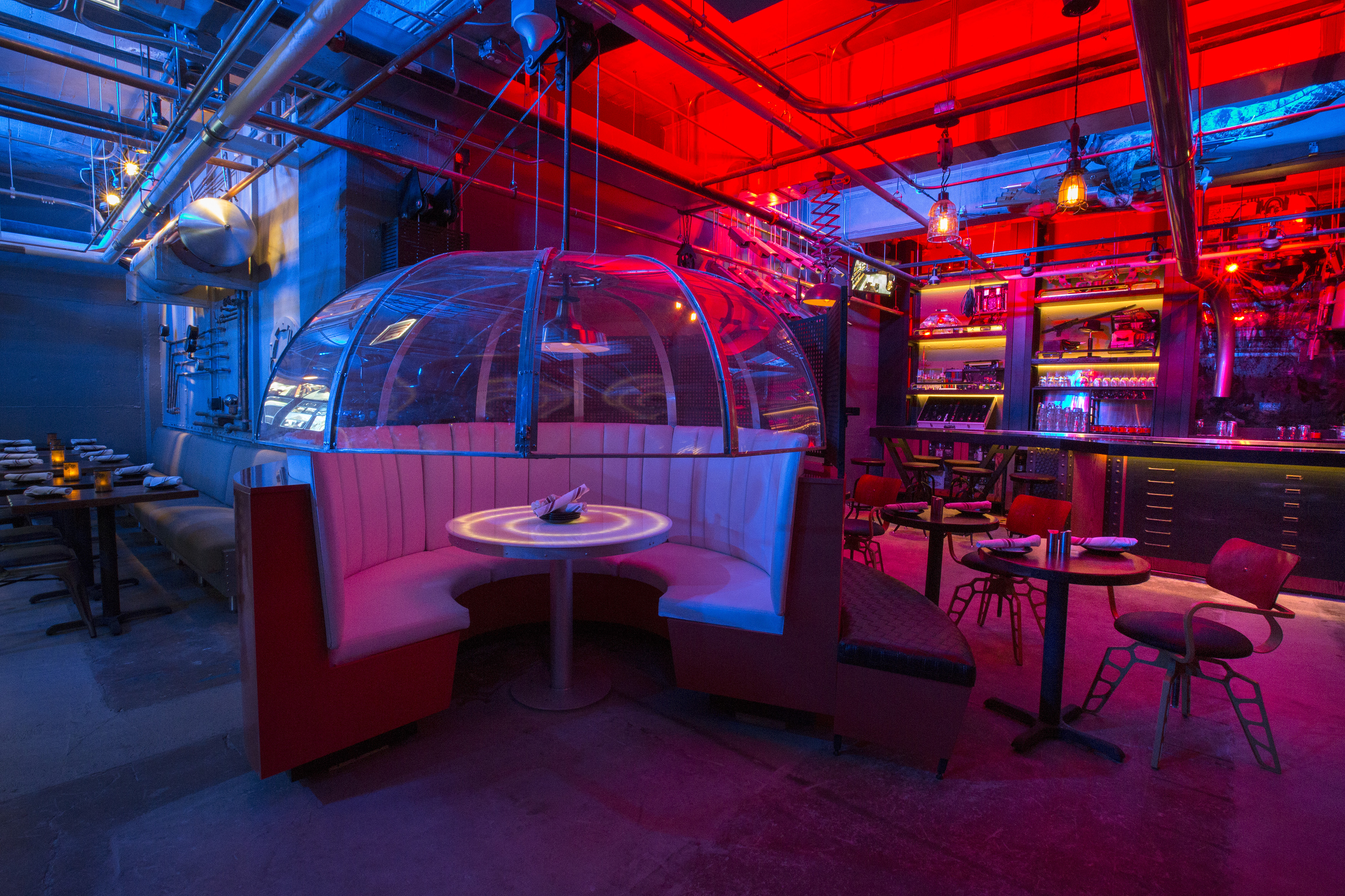 A red- and blue-lit industrial dining room with a booth covered by a clear umbrella-shaped dome.