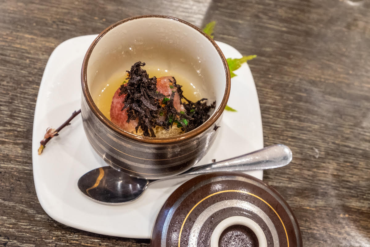 A serving cup on a platter with a spoon holds black truffle grilled rice with parmesan dashi and wagyu lardon.