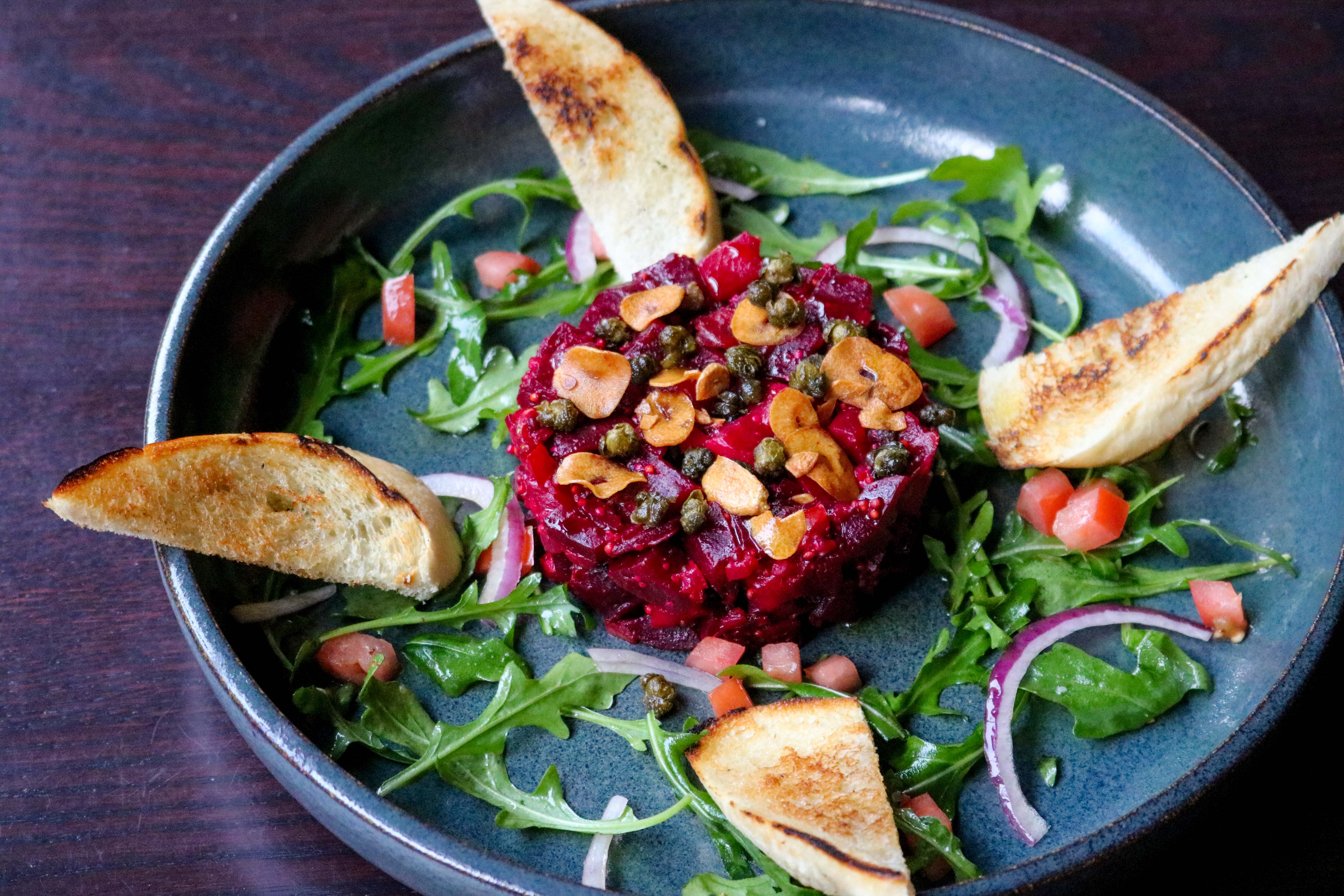 A dish of red beet tartare encircled with four slices of grilled bread in a shallow bowl.