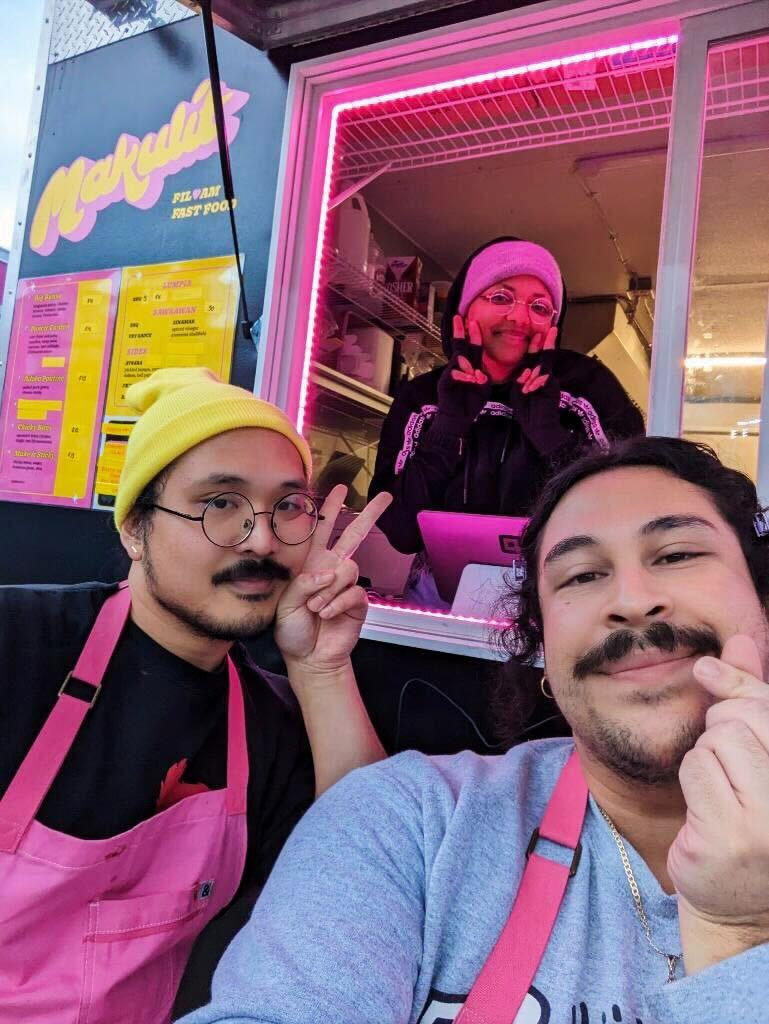 Three people posing at the window of a food cart.