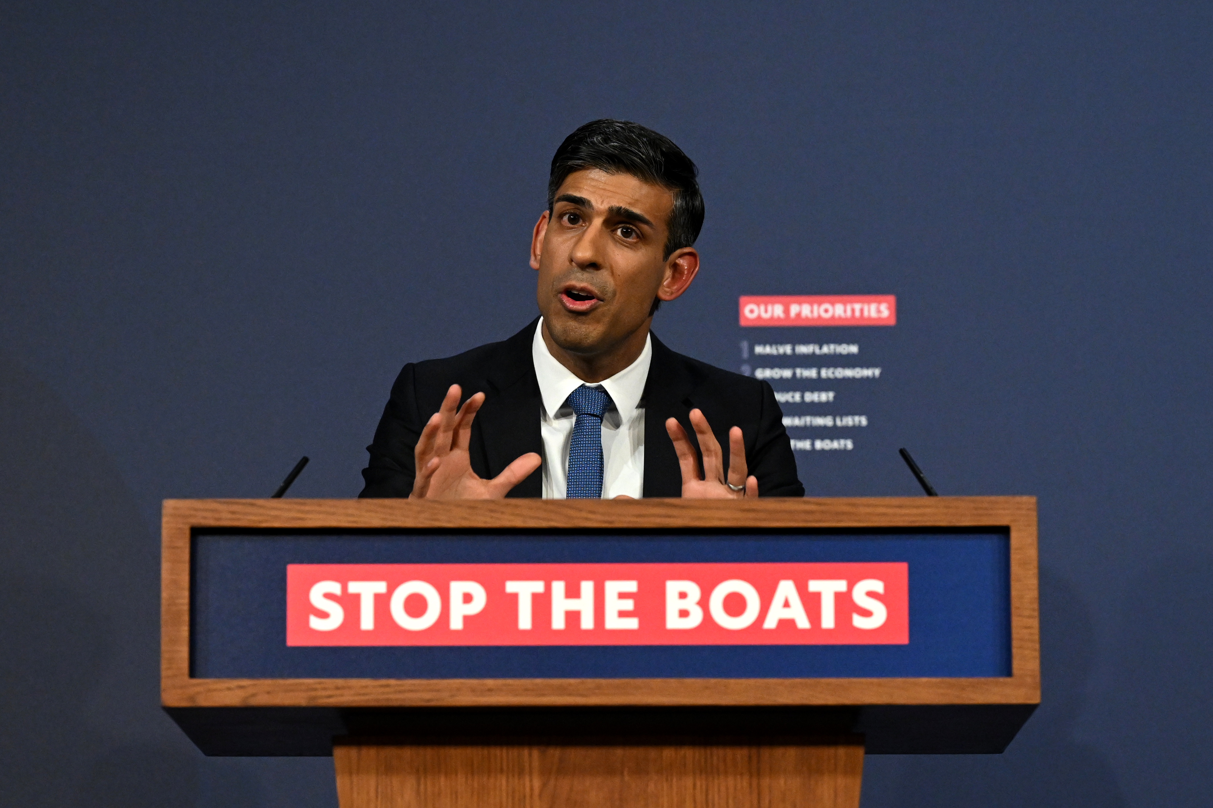 British Prime Minister Rishi Sunak, wearing a dark suit with a white shirt and blue tie, gestures while speaking from behind a podium with a sign reading “Stop the Boats.”
