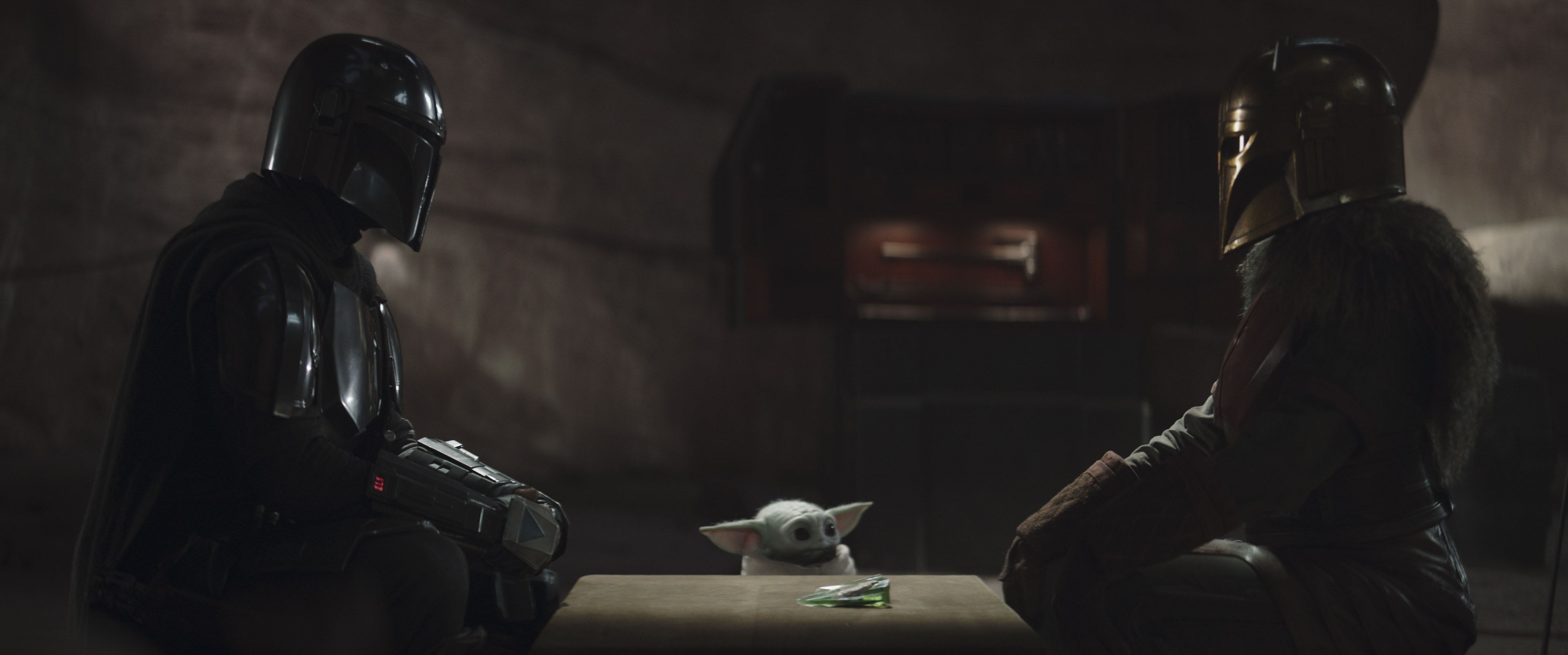 The Mandalorian (Pedro Pascal) sitting and looking at the Armorer (Emily Swallow) with Grogu (Baby Yoda) in between