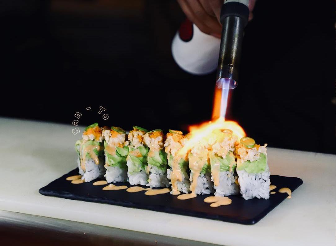 A flame crusts the top of a cut hand roll on a slate plate.