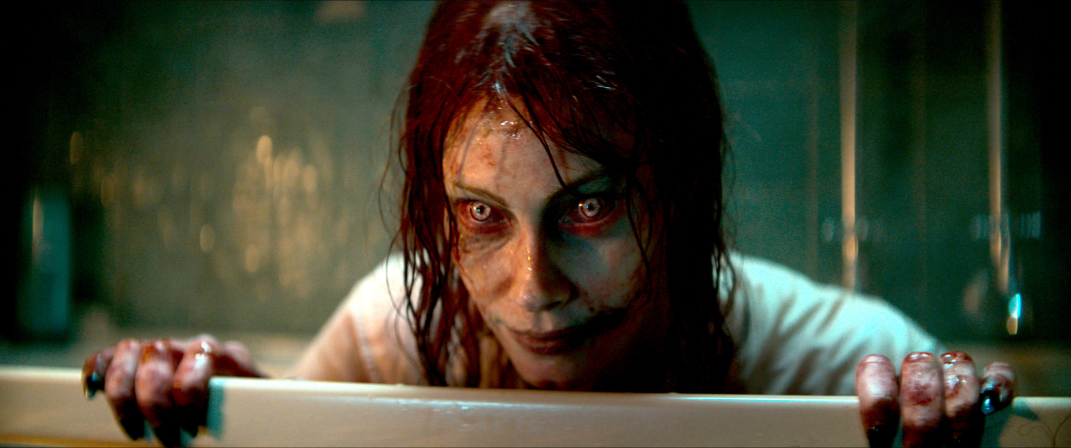 Ellie (Alyssa Sutherland), possessed and turned into a red-and-yellow-eyed, greasy-haired, grimy-faced Deadite, smiles eerily over a barrier in Evil Dead Rise