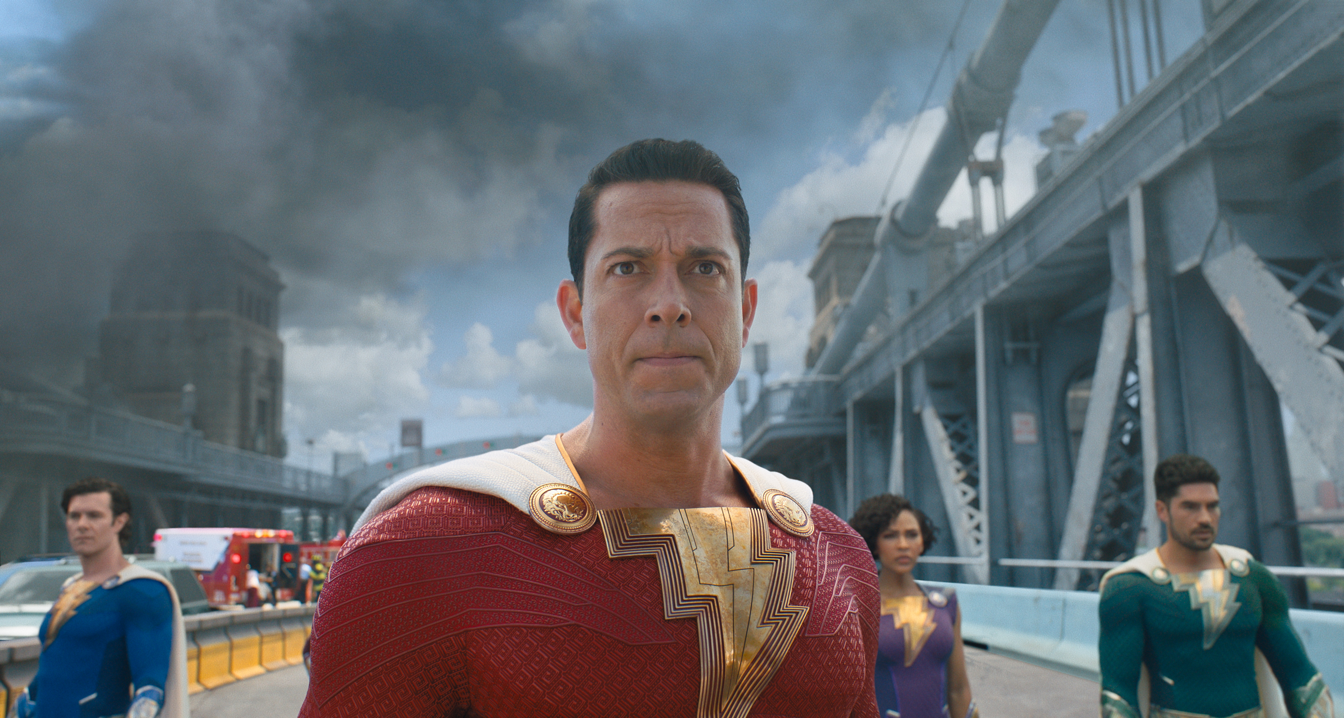 A superhero in a red suit is flanked by three other heroes in blue suits, all on a busy city street alongside a bridge, with thunderclouds above them.