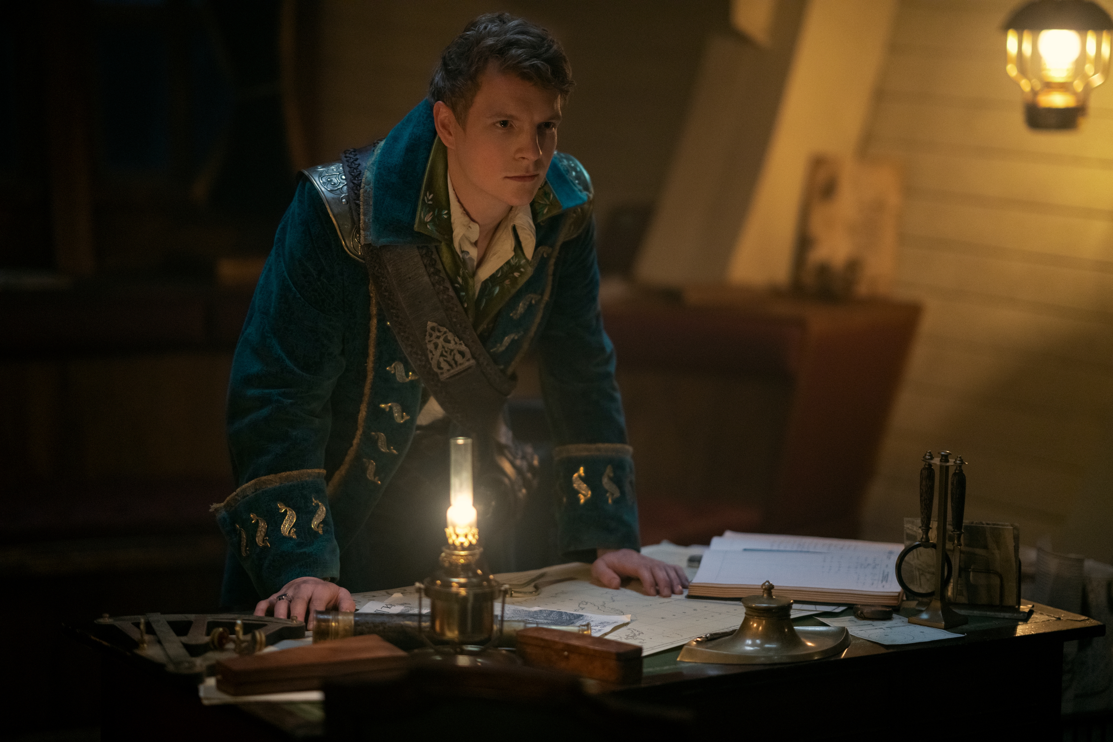 In a a still from Shadow and Bone season 2, Patrick Gibson as Sturmhond leans forward with his hands resting on a cluttered desk in his ship’s cabin.