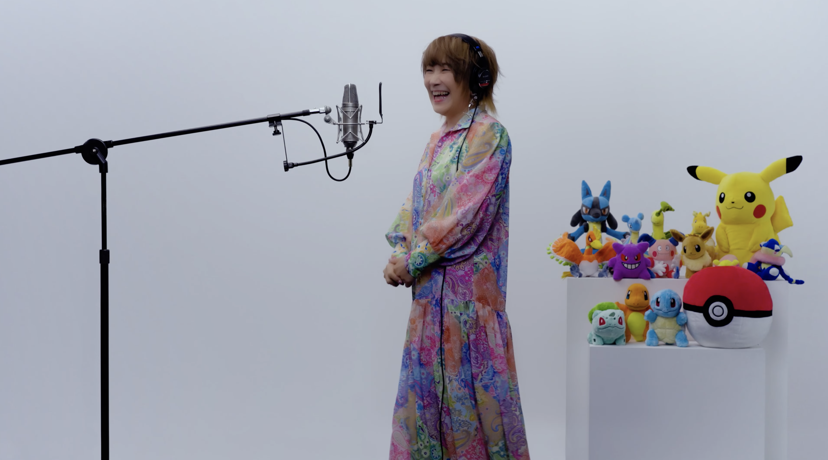 Rika Matsumoto signing on a bare white set. She’s smiling. The only thing on set is a collection of Pokémon plushies like Pikachu, Squirtle, and the other starter  Pokémon.