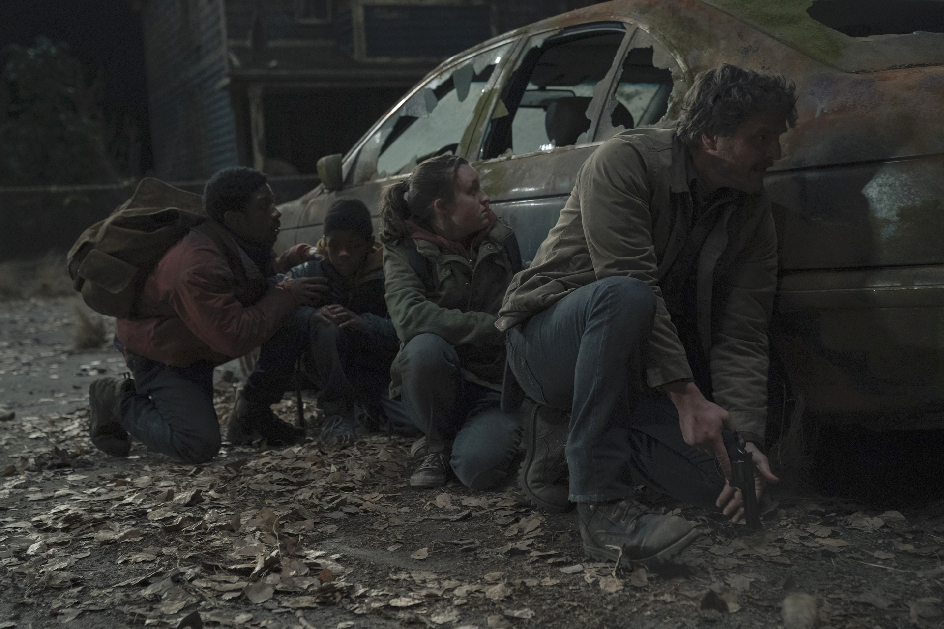 Joel (Pedro Pascal), Ellie (Bella Ramsey), Henry, and Sam crouching behind a car