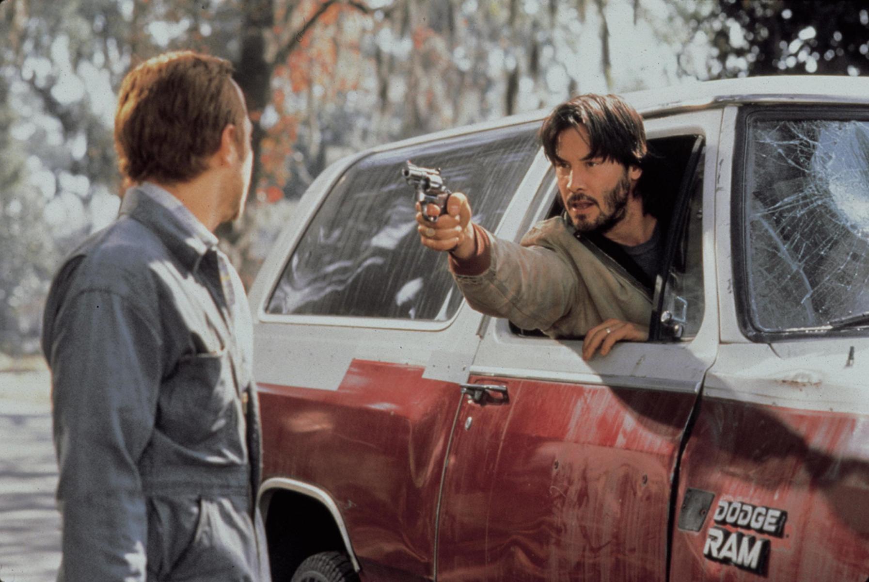 A bearded Keanu Reeves points a revolver at a man in The Gift. Reeves is sitting in the passenger seat of a white and red Dodge Ram, with his head and arm out of the window.