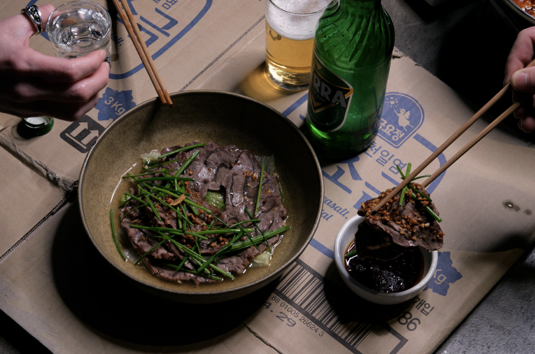 A hand holding chopsticks dunks a piece of cooked meat into a dipping sauce while another hand clutches a shot of clear alcohol.