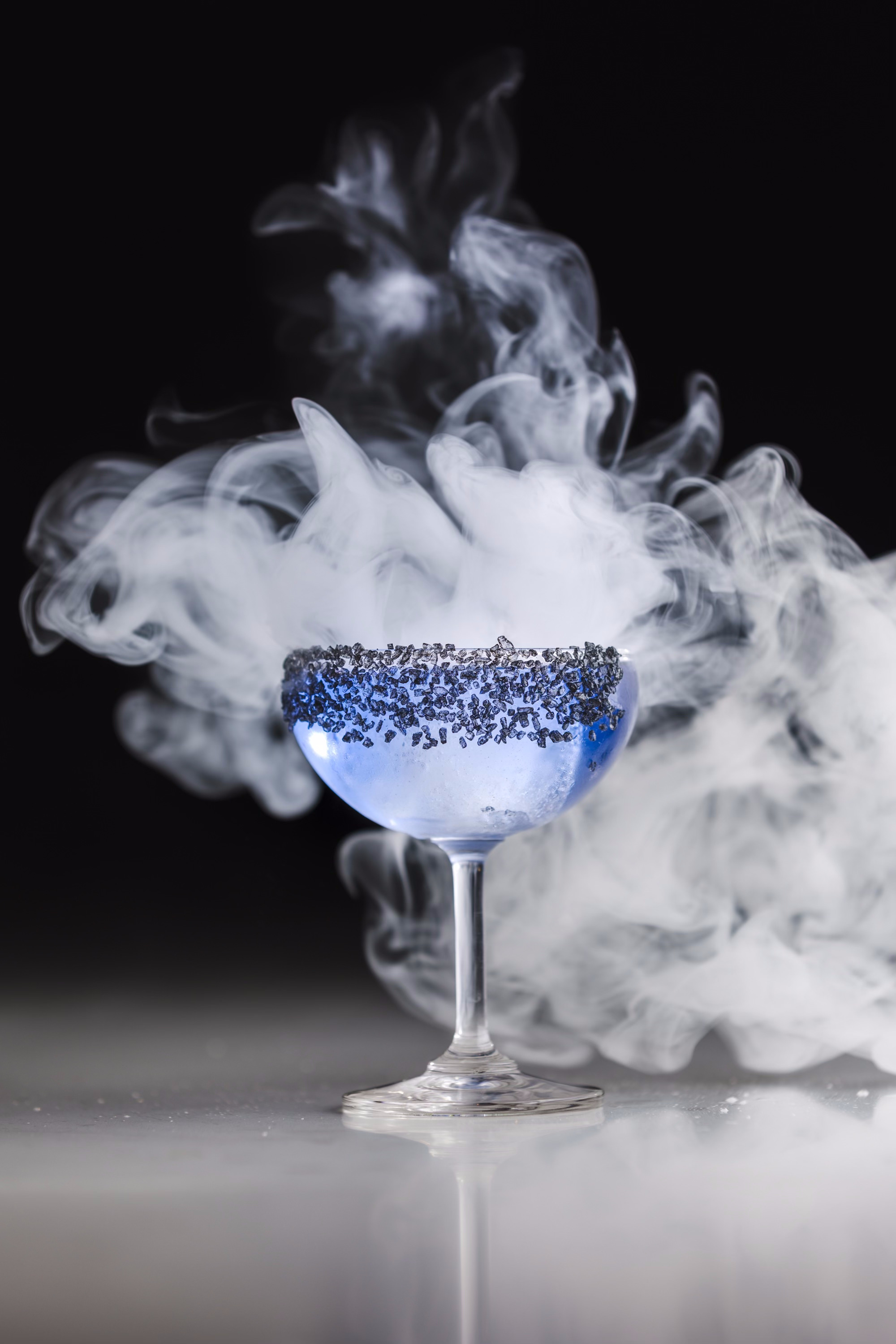 A blue-tinged cocktail with lavender around the rim and a cloud of white smoke behind it.