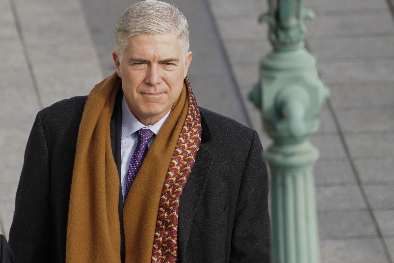 Justice Gorsuch is walking outside on a sunny, cold day, wearing a suit, a heavy overcoat, and a gold-and-red patterned scarf.