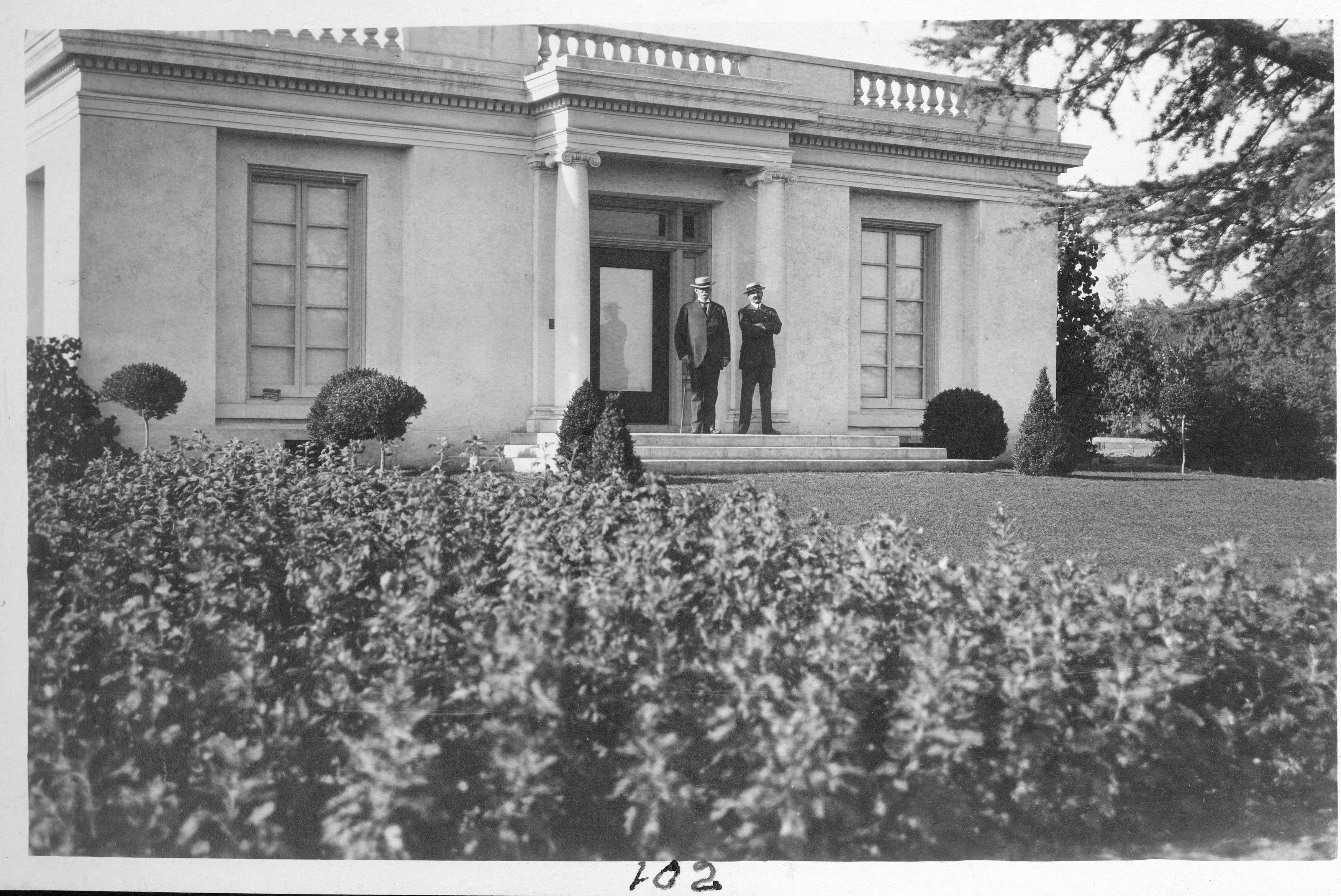 A black and white historic photograph showing two men standing in front of a white building.