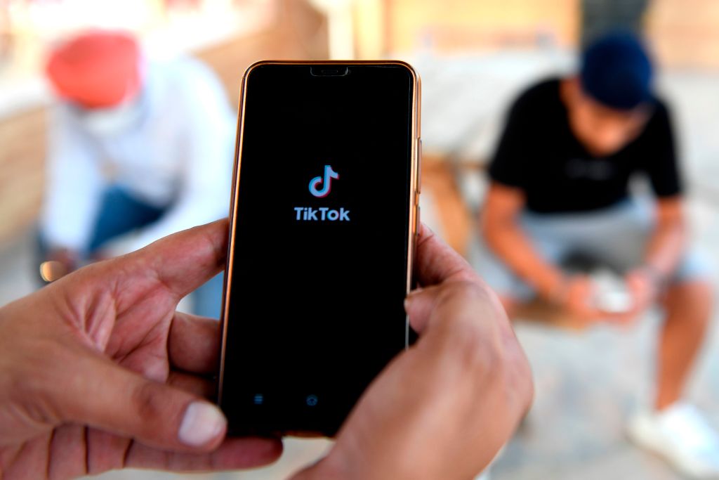 A person’s hands hold a smartphone with the TikTok app open on it. In the background, two other people are looking at their phones.