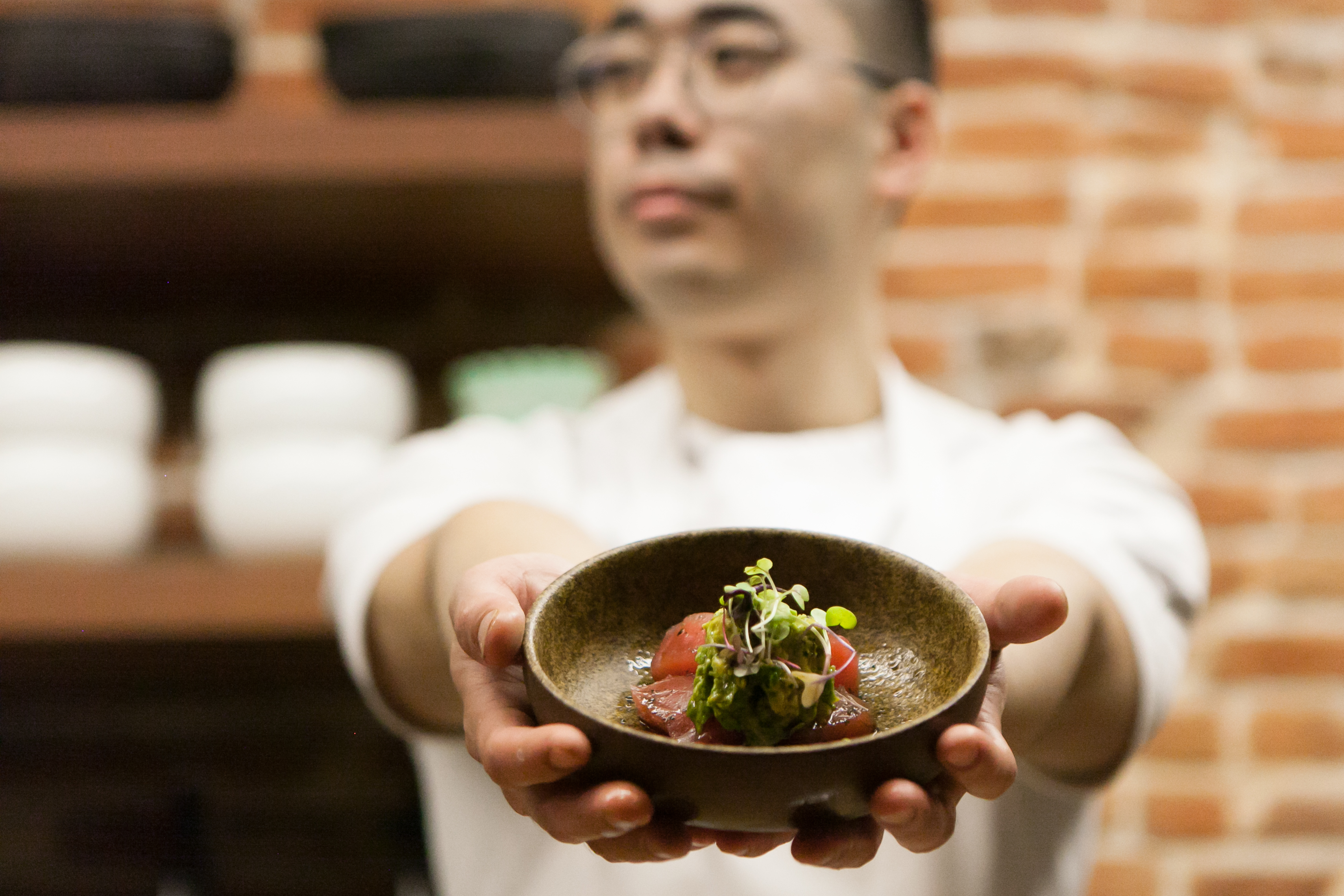 A male chef of Asian descent with arms outstretched in front of him holds a plate of sushi cupped in both hands while he looks away from the camera
