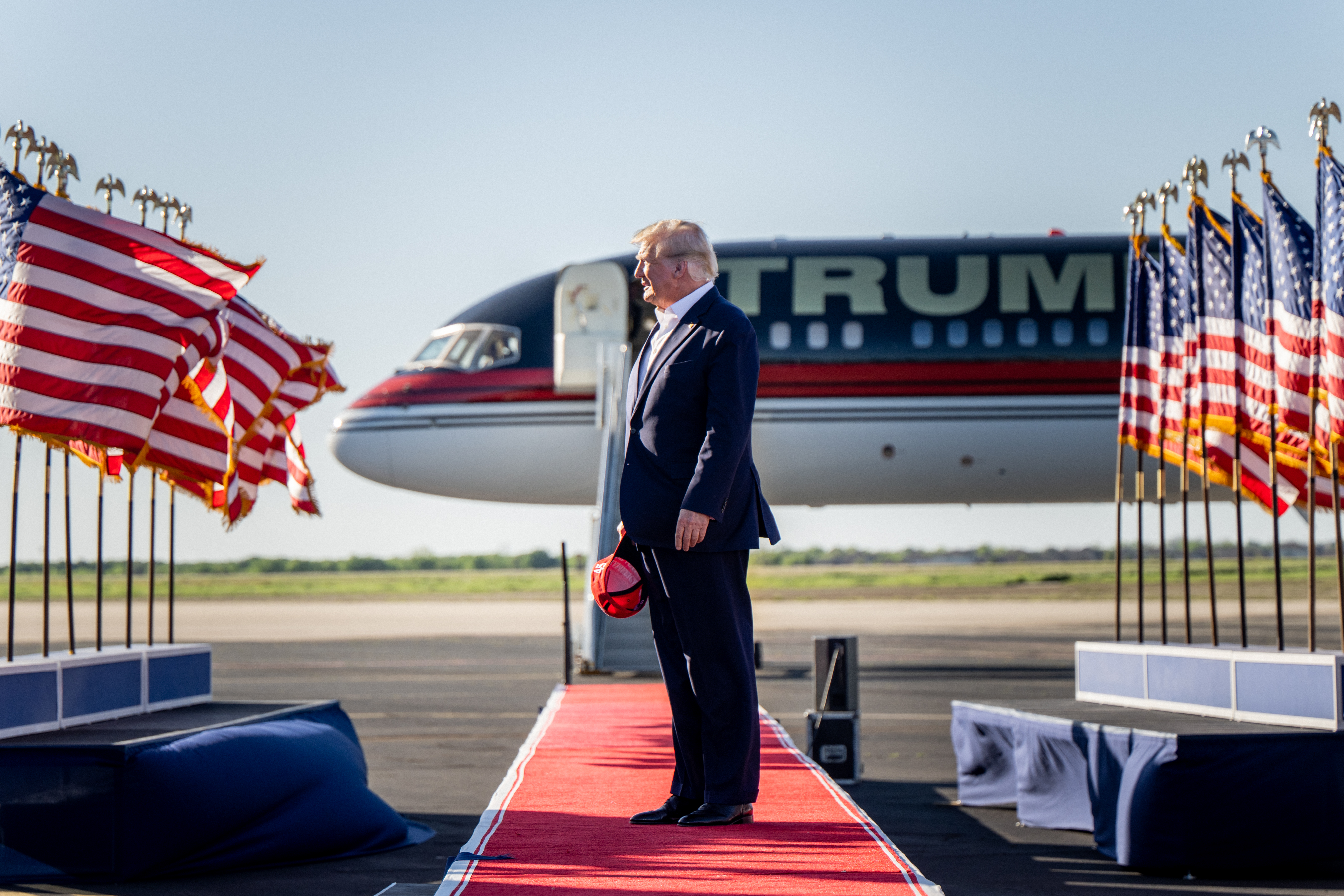 Trump tieless in a navy suit and white shirt, is seen in profile, standing between rows of flapping US flags, his plane, reading TRUMP behind him. He’s looking down, his MAGA hat in his hand.