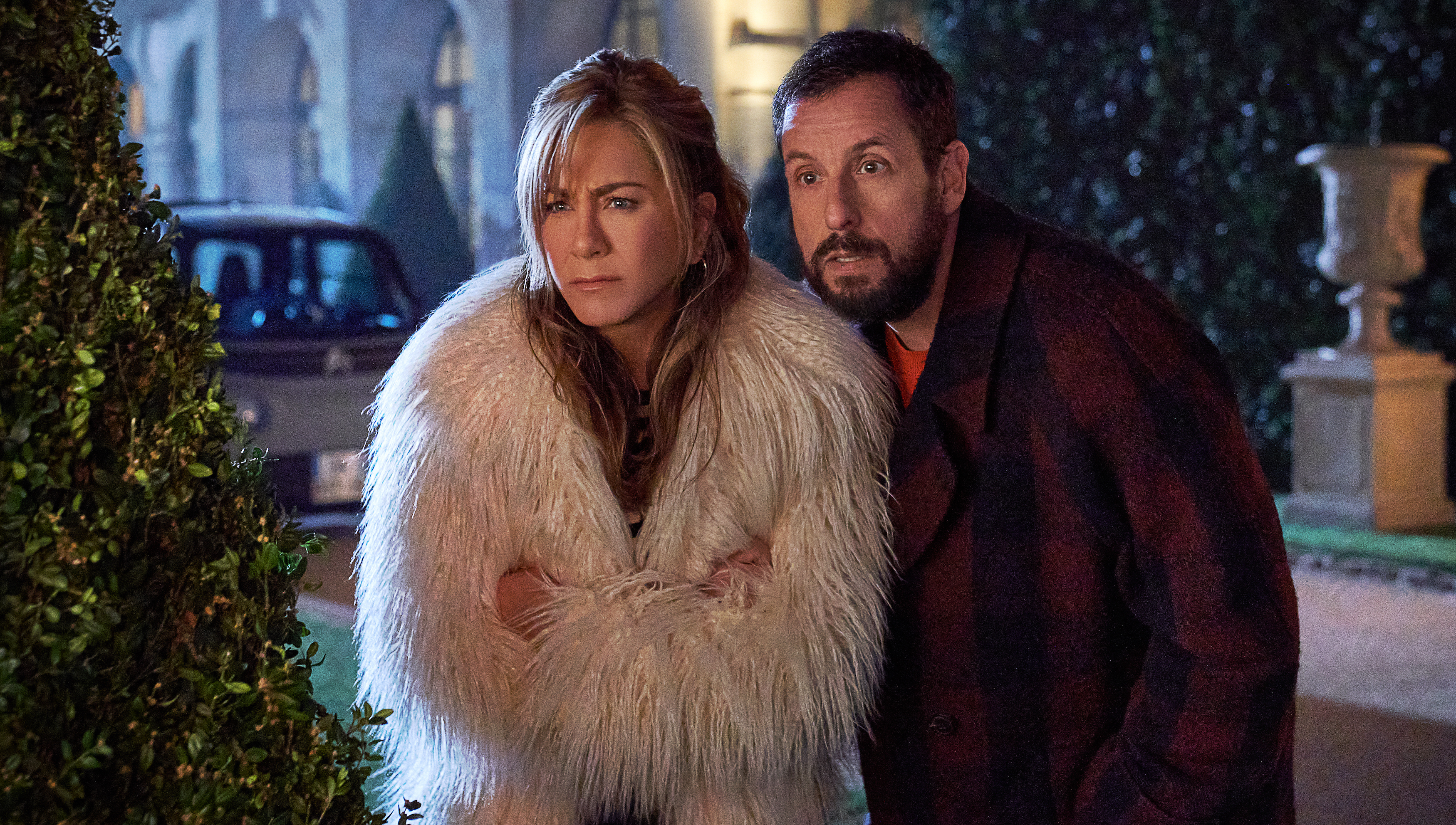 Audrey (Jennifer Aniston, in a giant fluffy white fur coat) glares and her husband Nick (Adam Sandler) boggles at something offscreen in a night shot in Netflix’s Murder Mystery 2