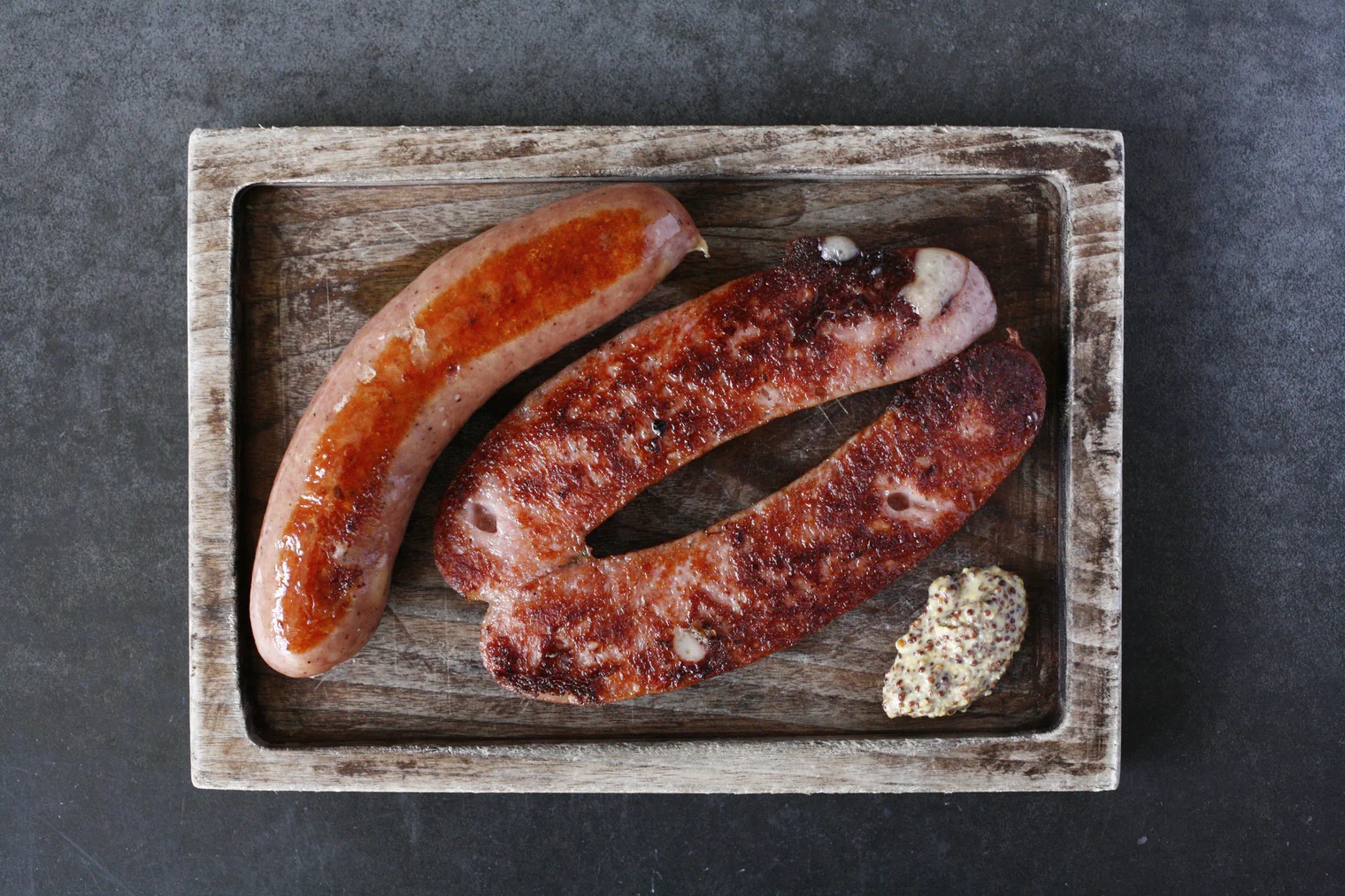 A wood platter of three half sausage links, pan-fried and browned, next to a dollop of grainy mustard.