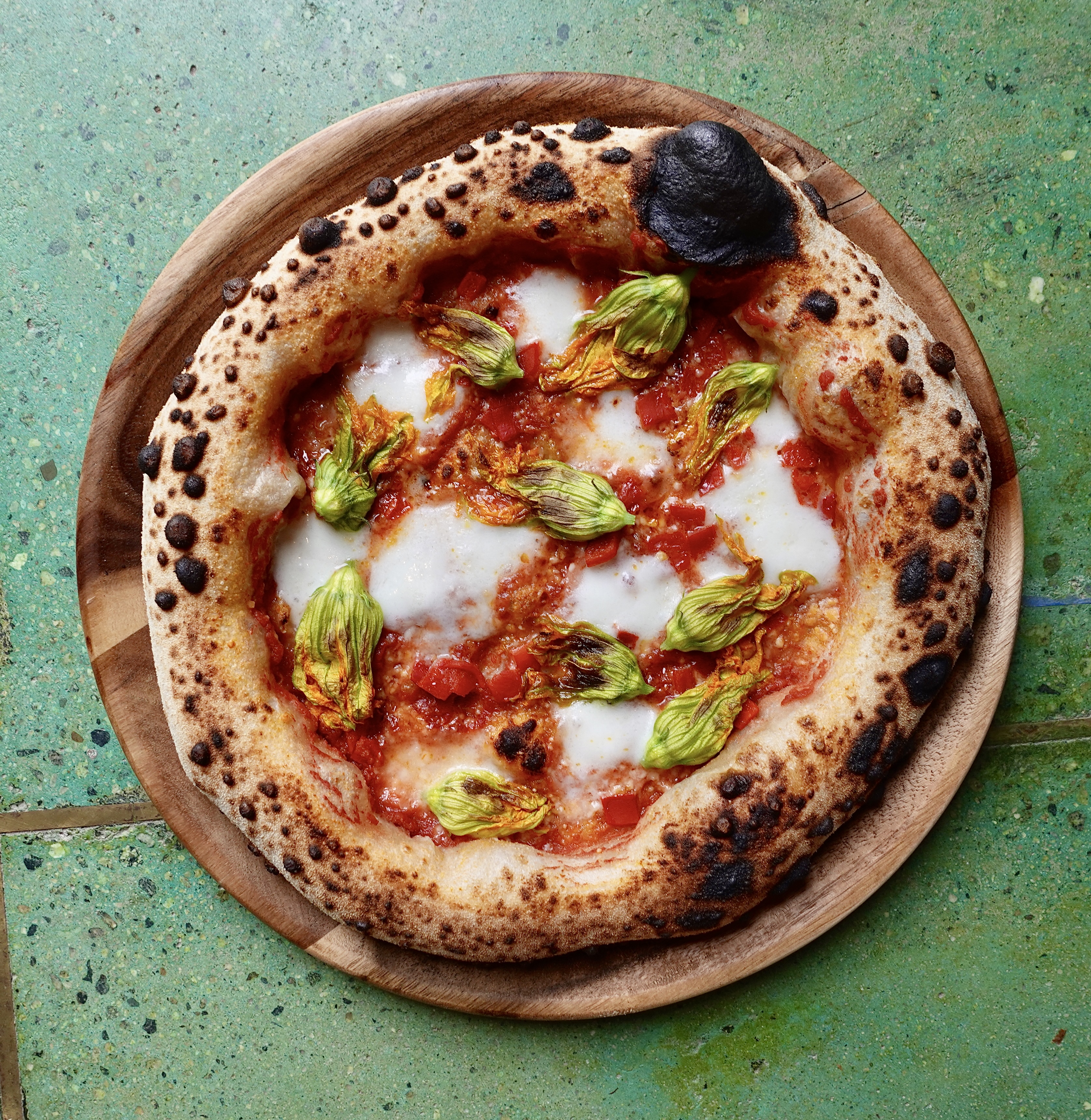 An overhead shot of a light green table with blistered pizza on top of a wooden plate, with ricotta and greens.