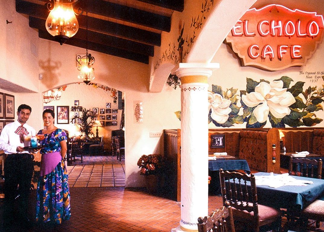 Two people in traditional Mexican outfits stand inside a bright restaurant with a neon sign.