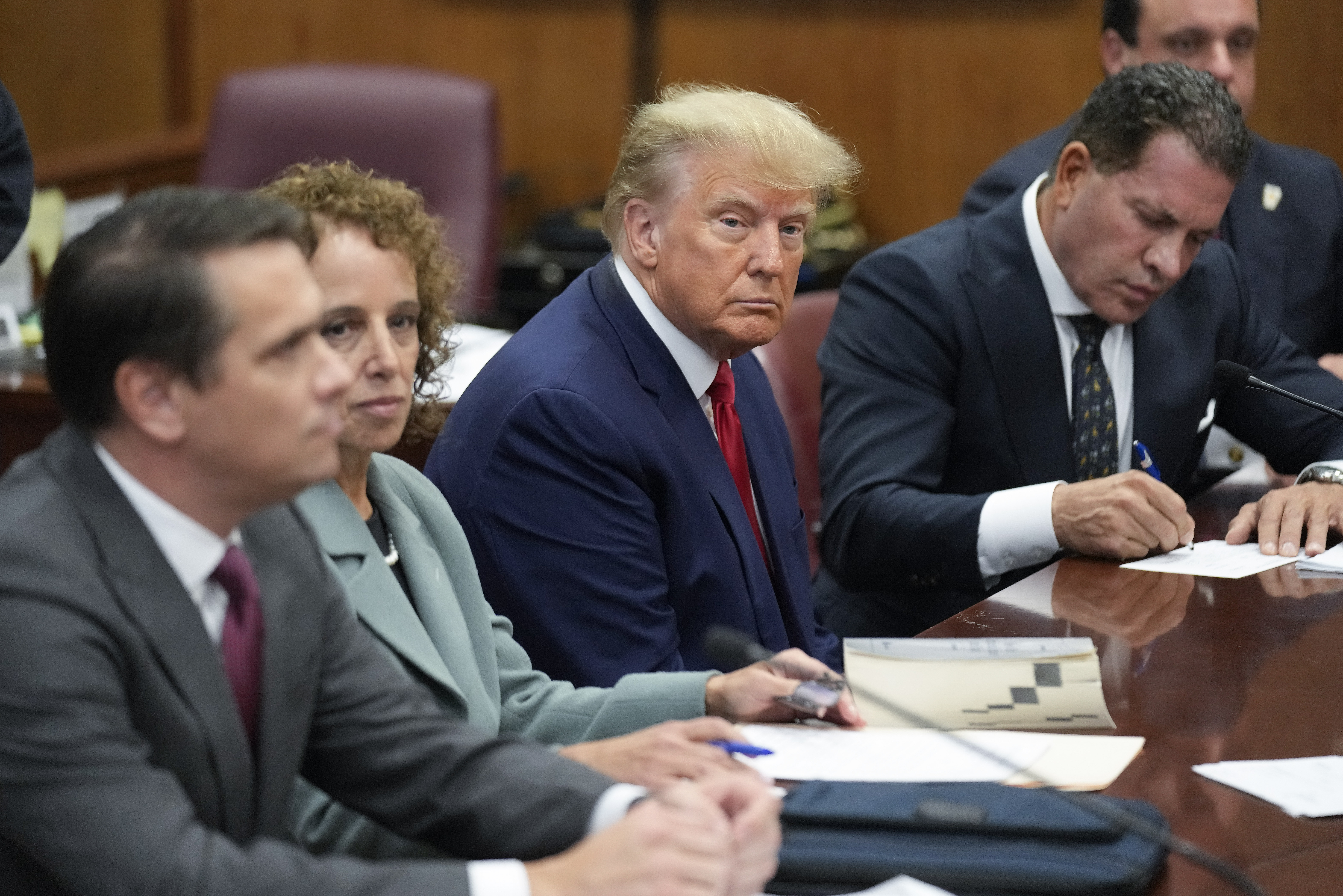 Trump in a blue suit, white shirt, and red tie, sits among a cluster of dark-suited lawyers, looking sternly at the photographer.