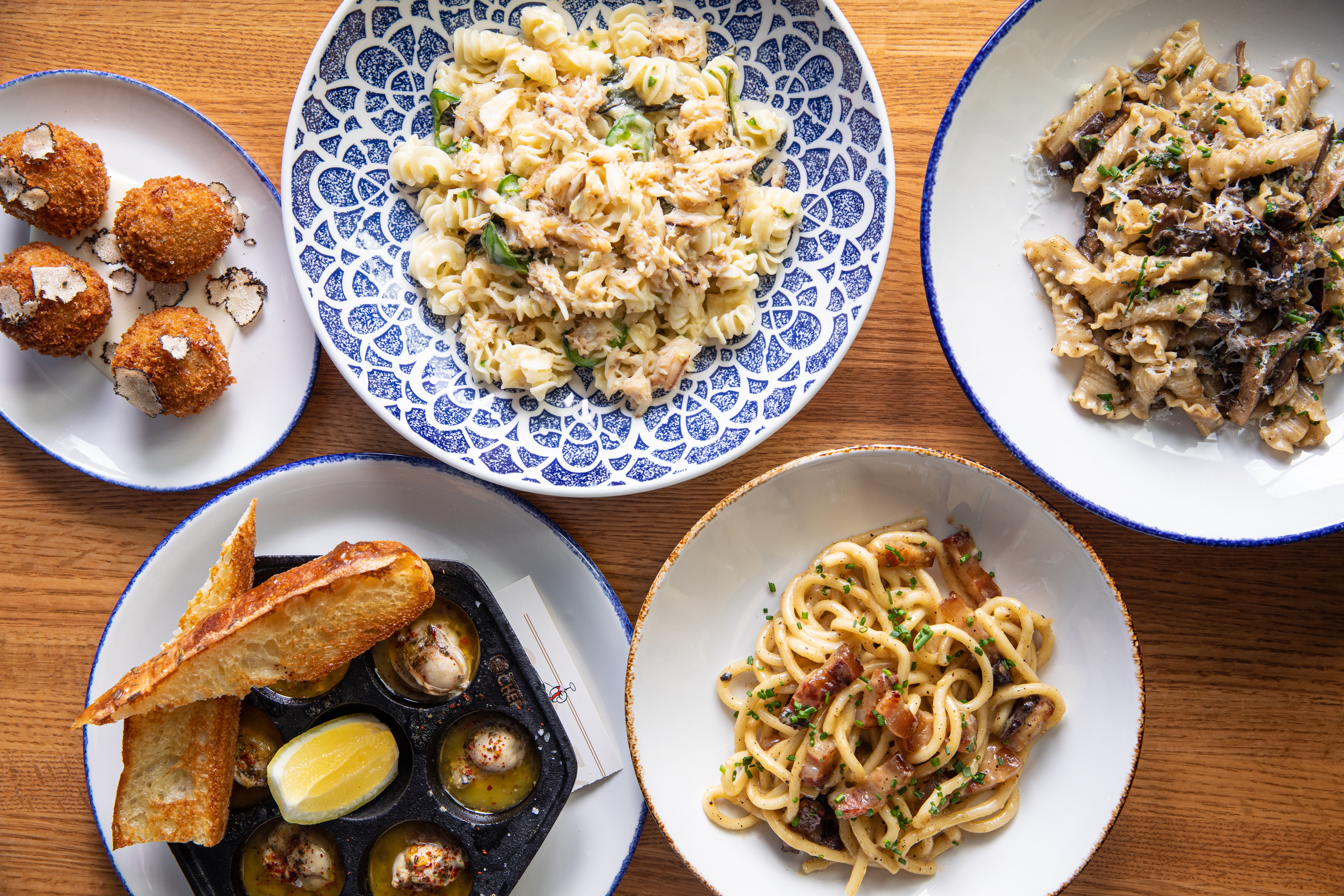 Overhead view of plates of arancini, radiatori pasta, campanelle pasta, six oysters in a cast-iron oyster plate, and bigoli pasta.
