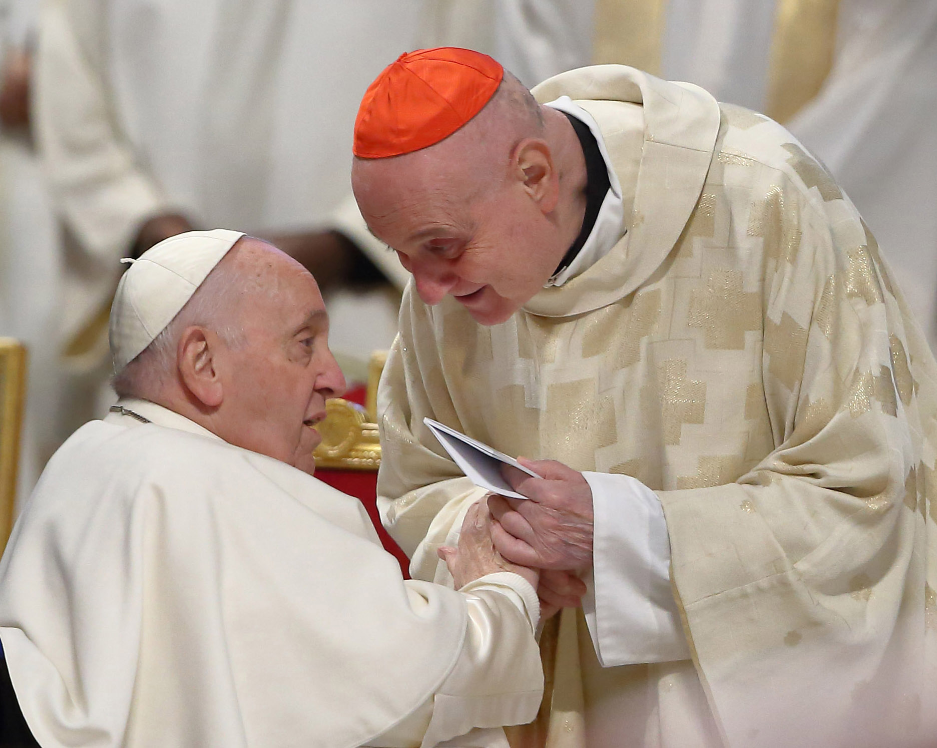 The Catholic pope, seated and wearing a white robe and cap, speaks with a bishop, standing and leaning in, wearing a white robe and red cap.
