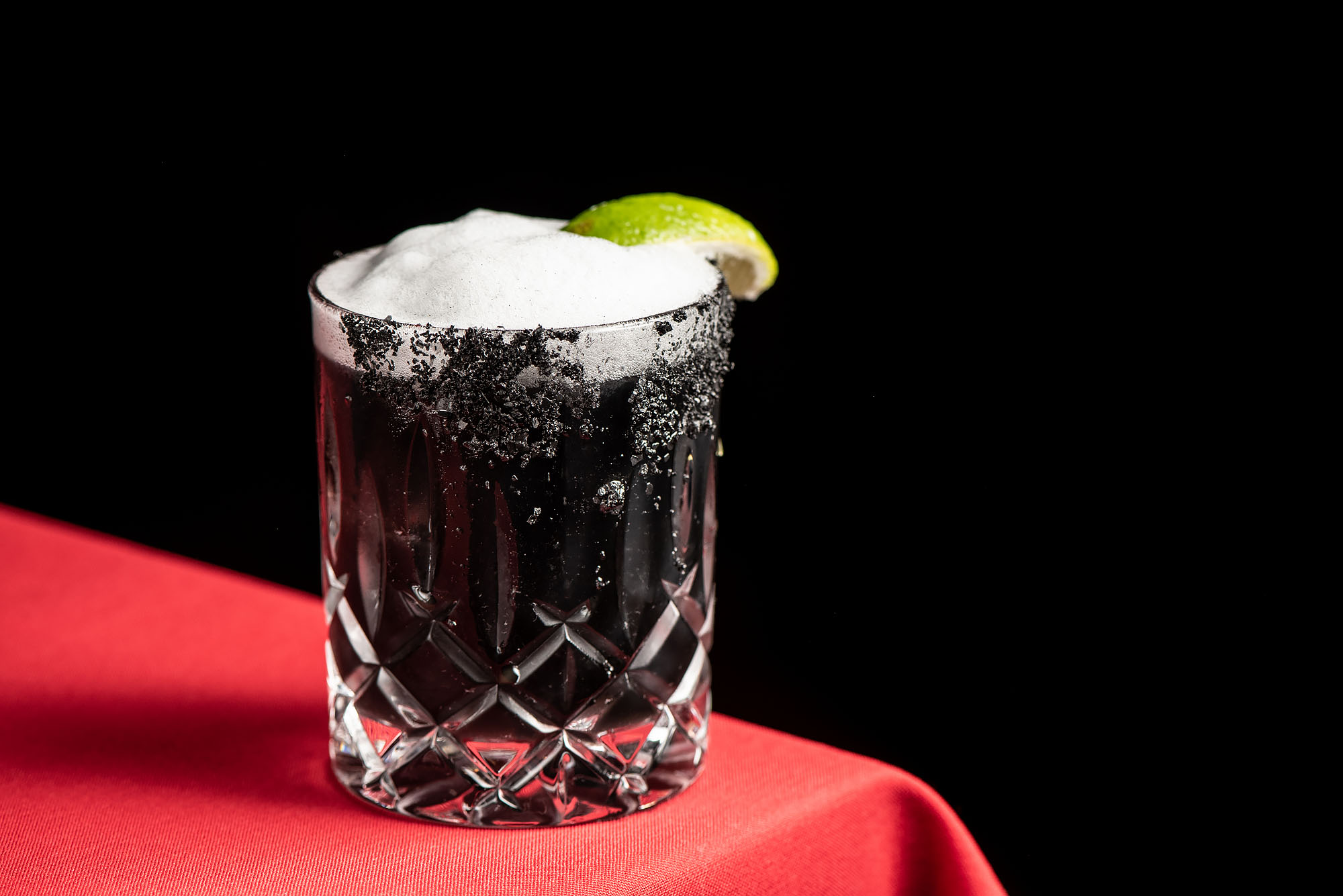 A black, fizzy cocktail with a lime wedge on a red tablecloth.