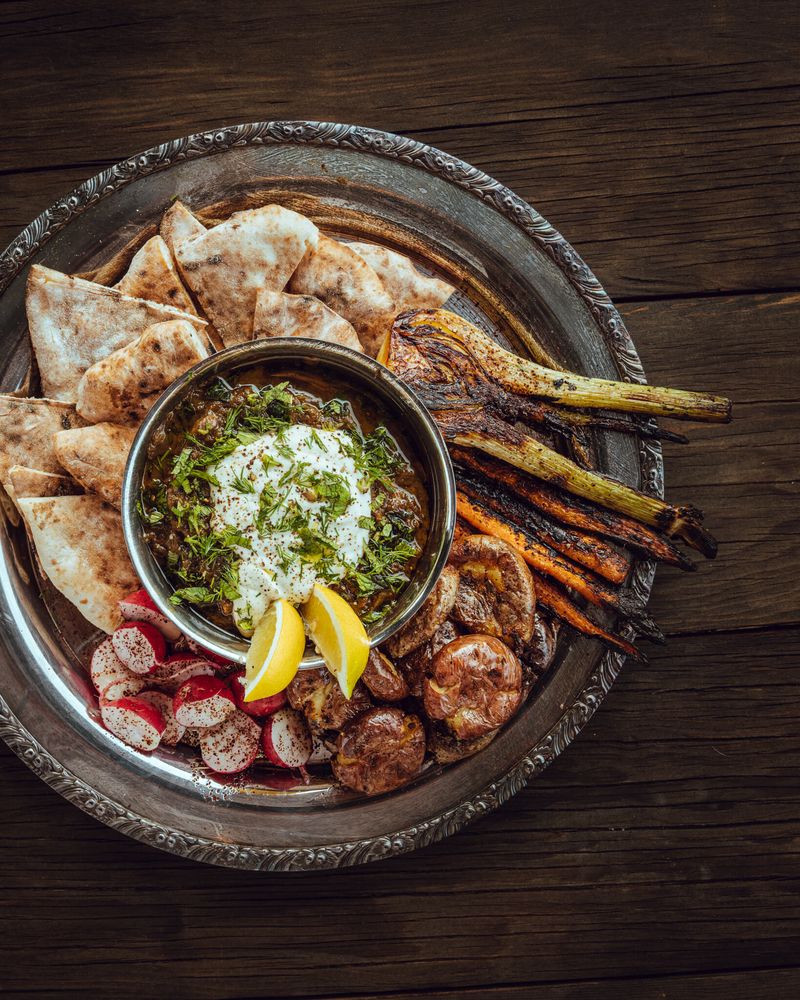 A pewter platter with a bowl of dip at the center surrounded by grilled veggies and pita.
