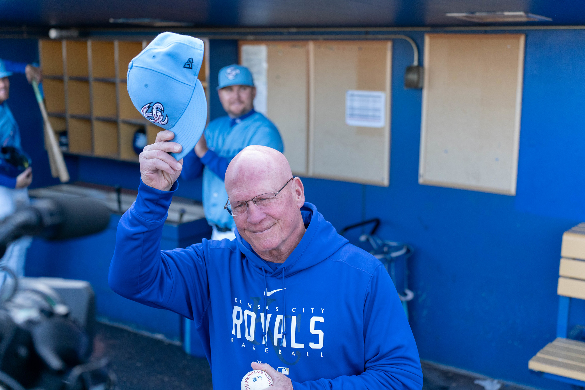 A view into the dugout of a white man with a bald head tipping his light blue baseball cap.