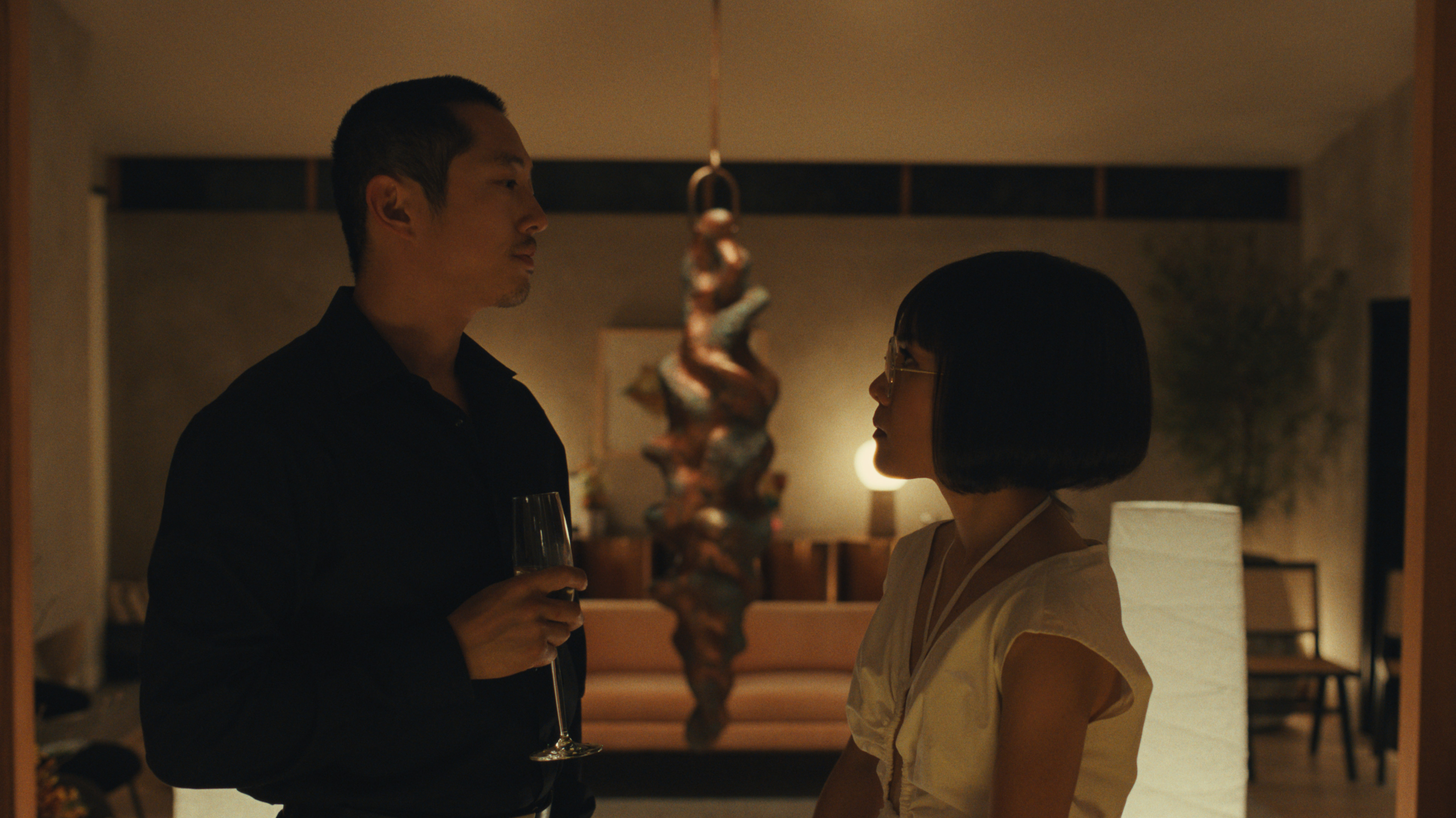 A taller Asian man holds a drink while looking at a shorter Asian woman wearing a classic bob wig.