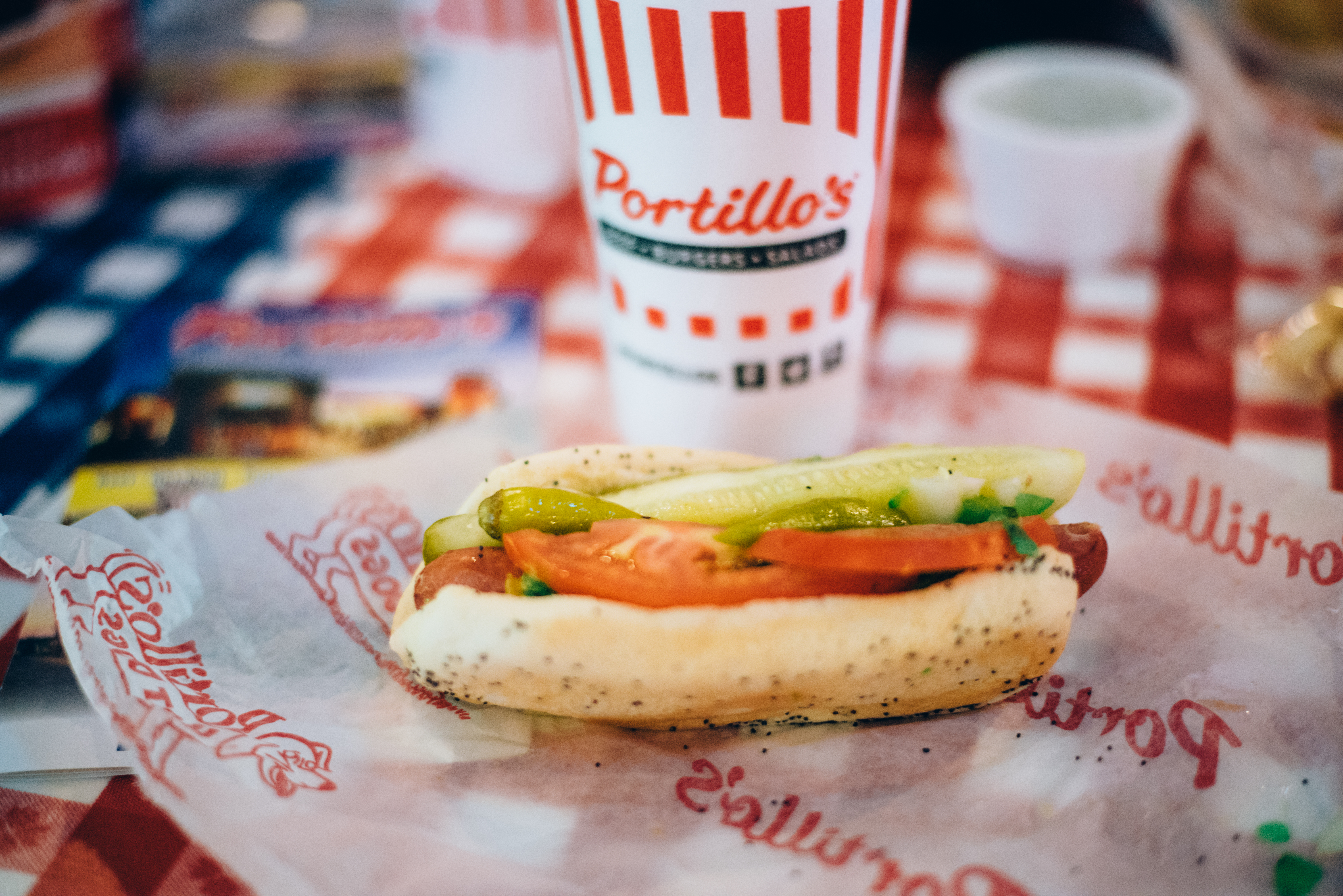 A Portillo’s Chicago-style hot dog on a wrapper with a fountain drink on a red checkered tablecloth.