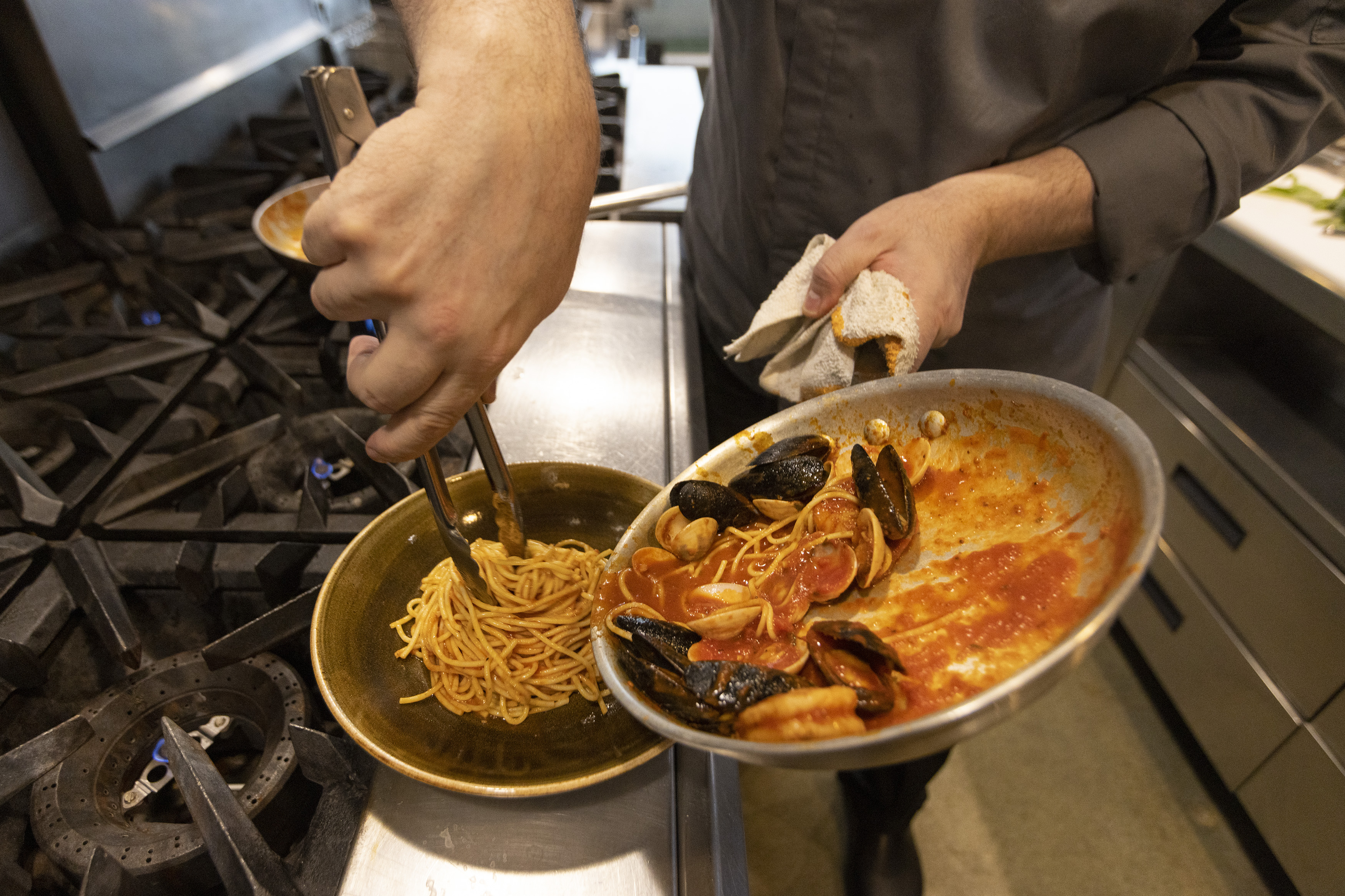 A chef uses tongs to plate a pasta dish in a restaurant kitchen.