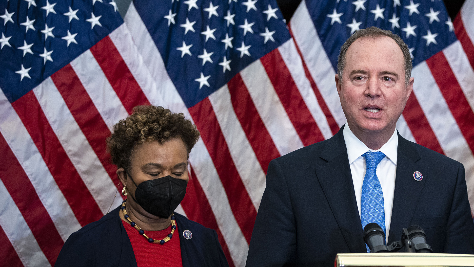 Schiff, a white man in a navy suit and light blue tie, speaks from a podium, while Lee, a Black woman in a black mask, black jacket and red shirt, stands next to him. Behind both is a large US flag.