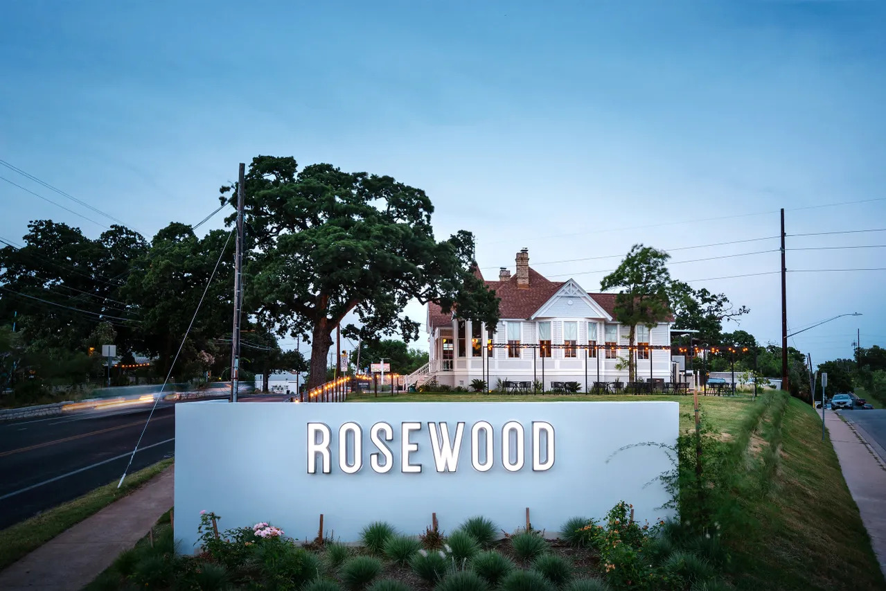 An outdoor sign that reads Rosewood in front of a lawn and building.