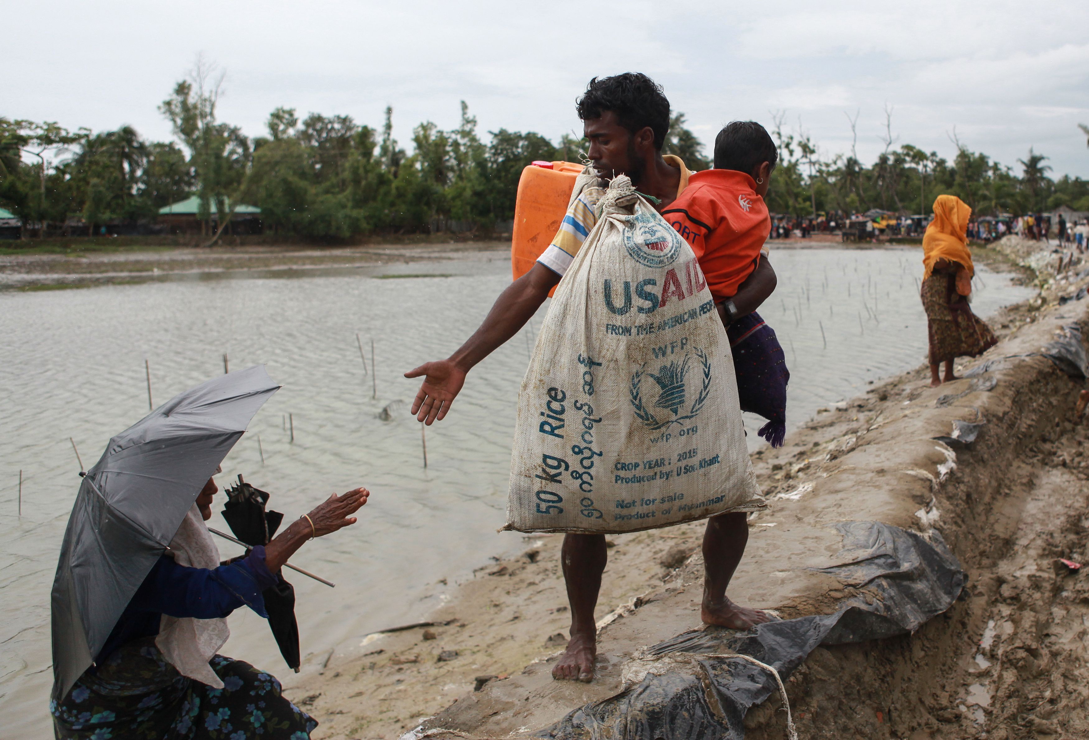 A man with an orange pack on his back and carrying a large white bag stamped with the letters USAID reaches down to give a woman holding an umbrella a hand up a riverbank.