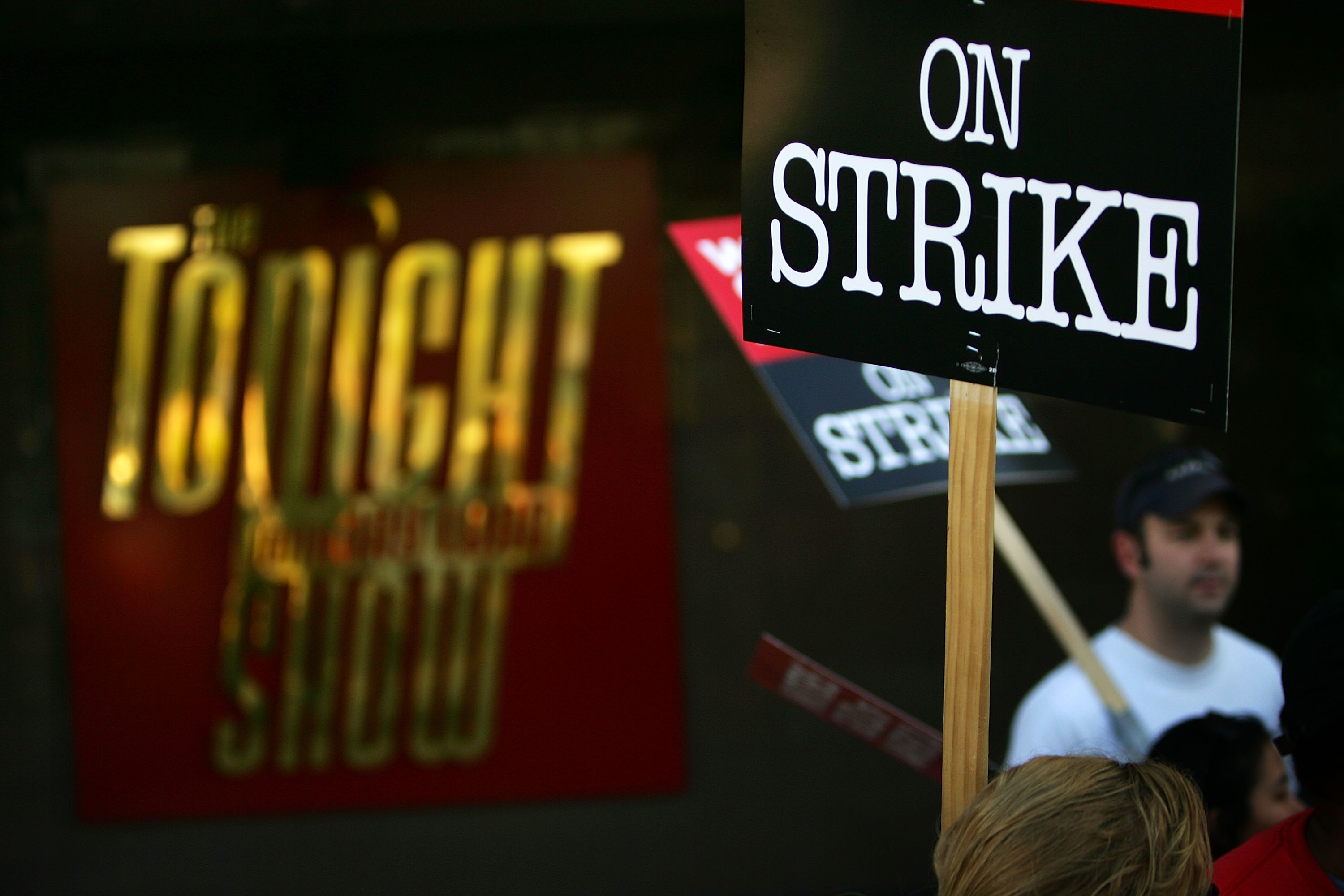 A man in a white shirt holds a sign reading “On strike” in front of a theater with the Tonight Show logo on it.