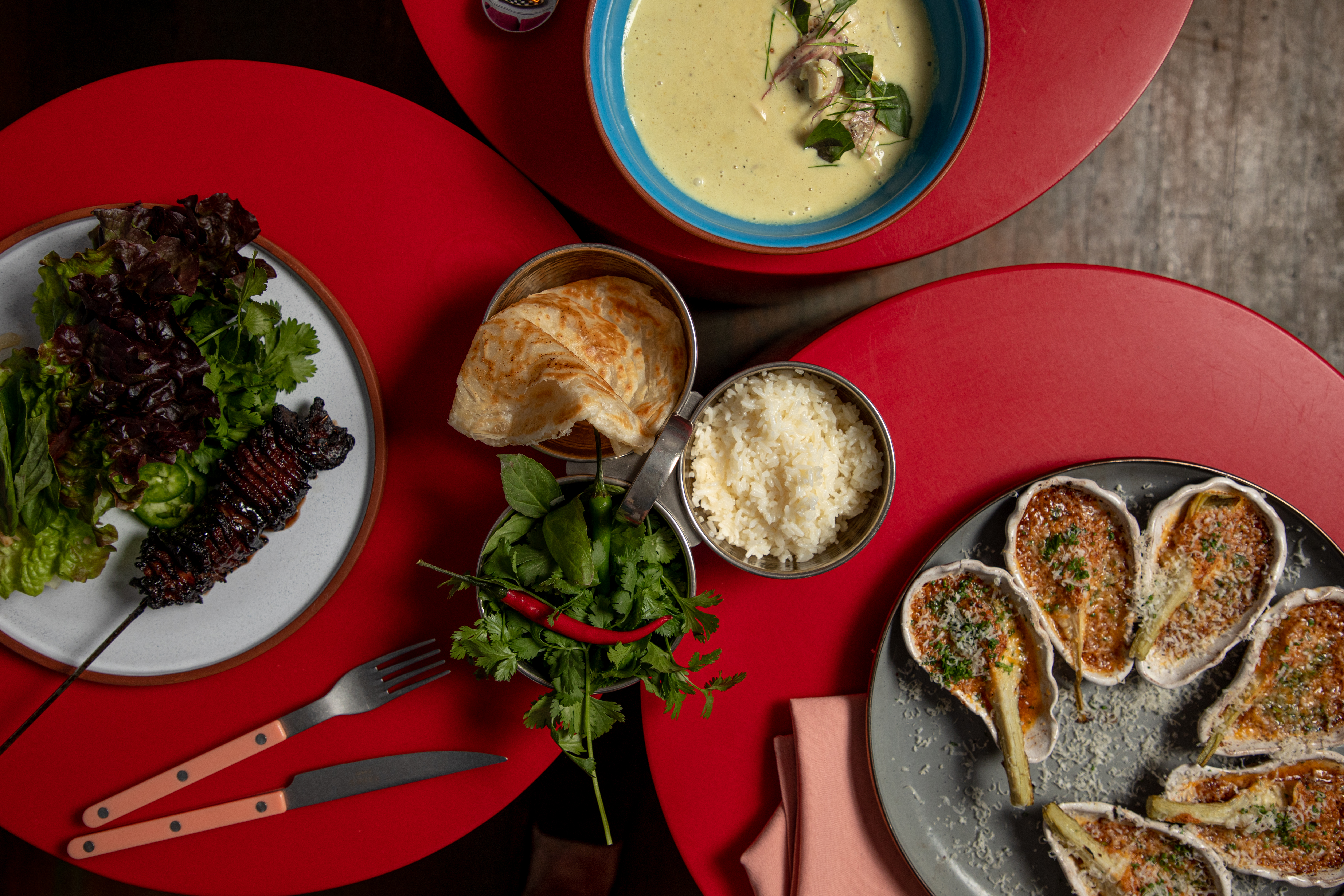An overhead shot of the dishes at Hungry Eyes, including oysters, roti, a meat skewer, and a soup
