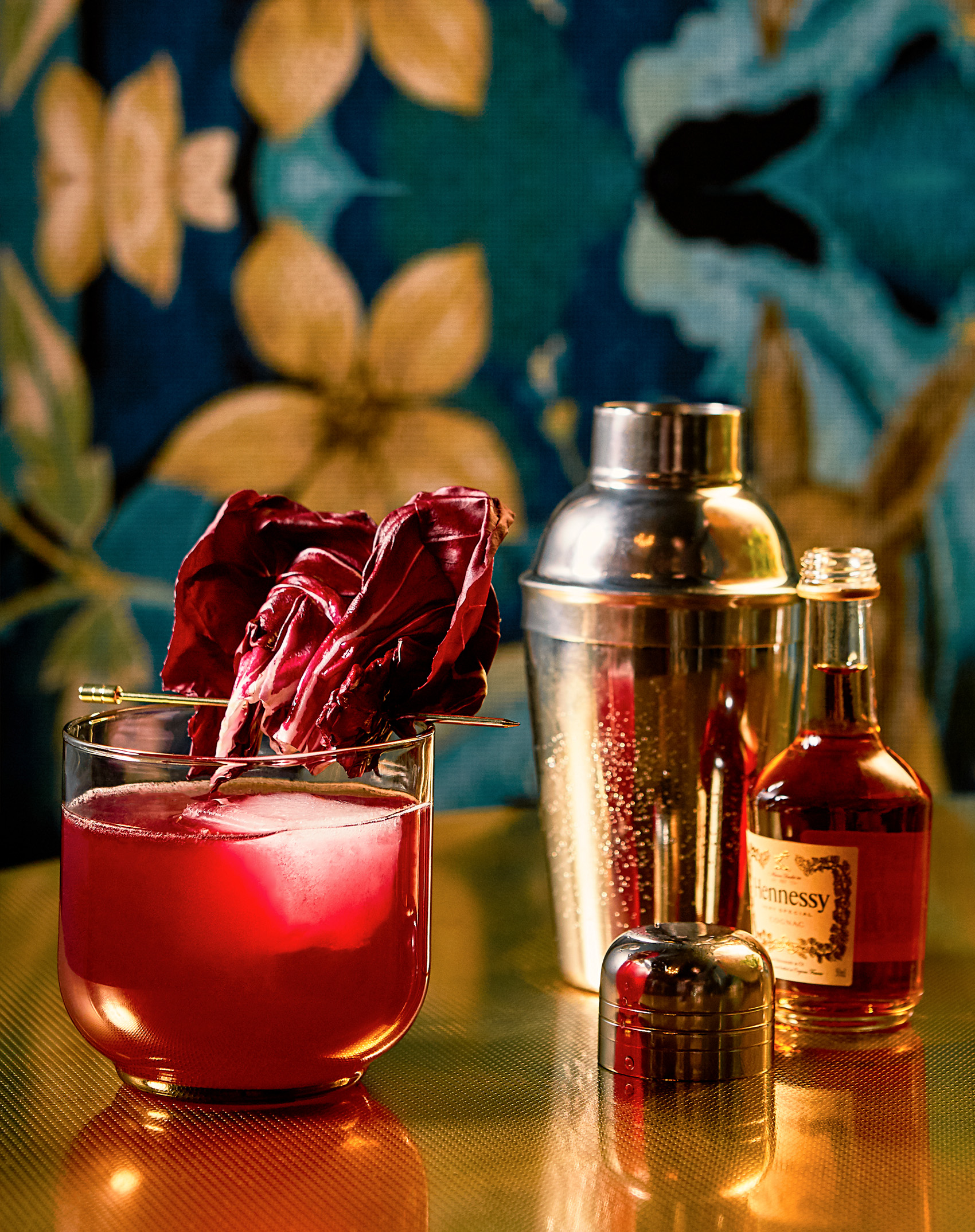 A red-hued cocktail in a stemless glass and garnished with pink flower petals. The glass sits on a dark wooden bar with mixers beside it, and floral wallpaper is visible in the background.