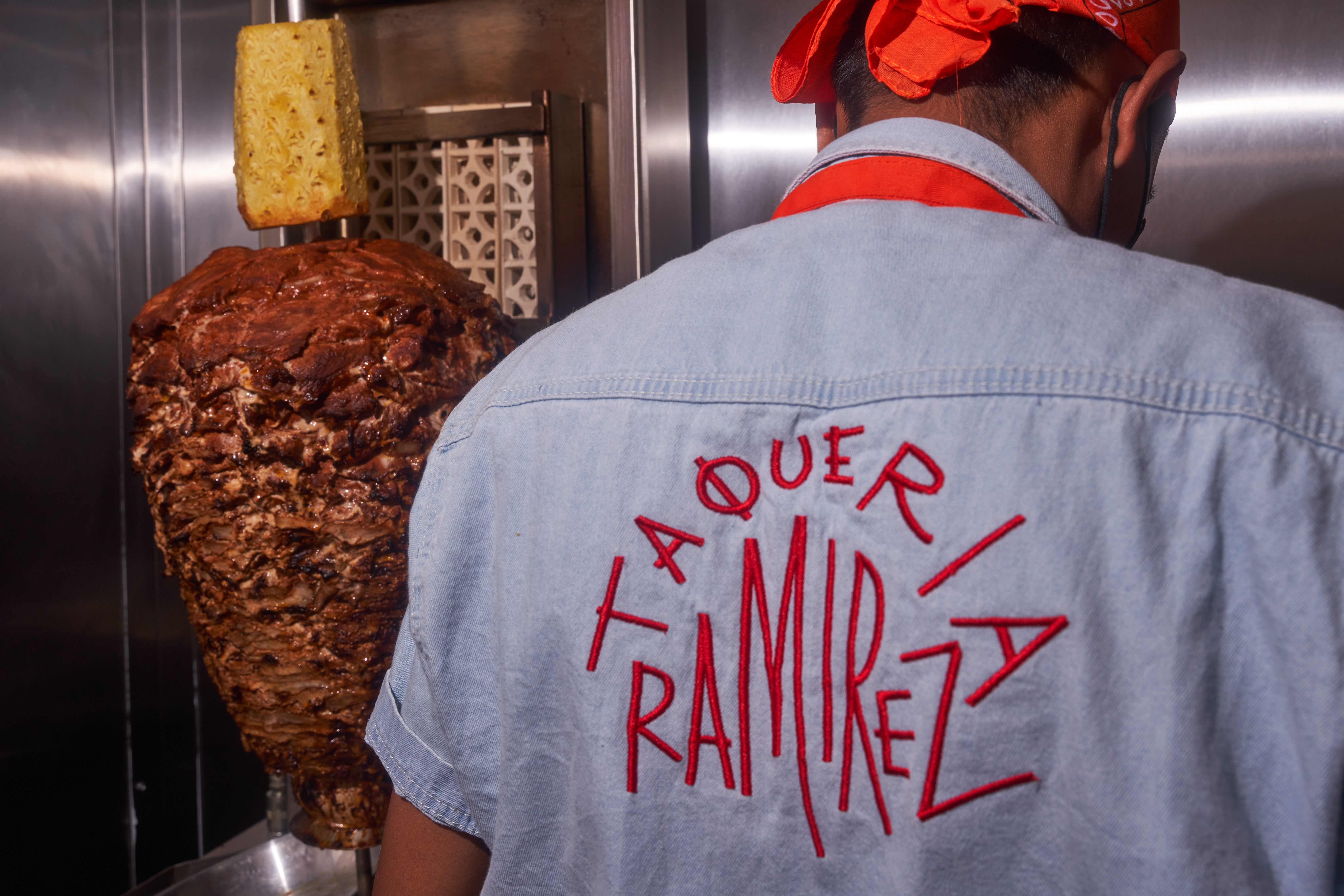 A man wearing a blue shirt with the words Taqueria Ramirez hand-stitched in red lettering stands behind an al pastor spit
