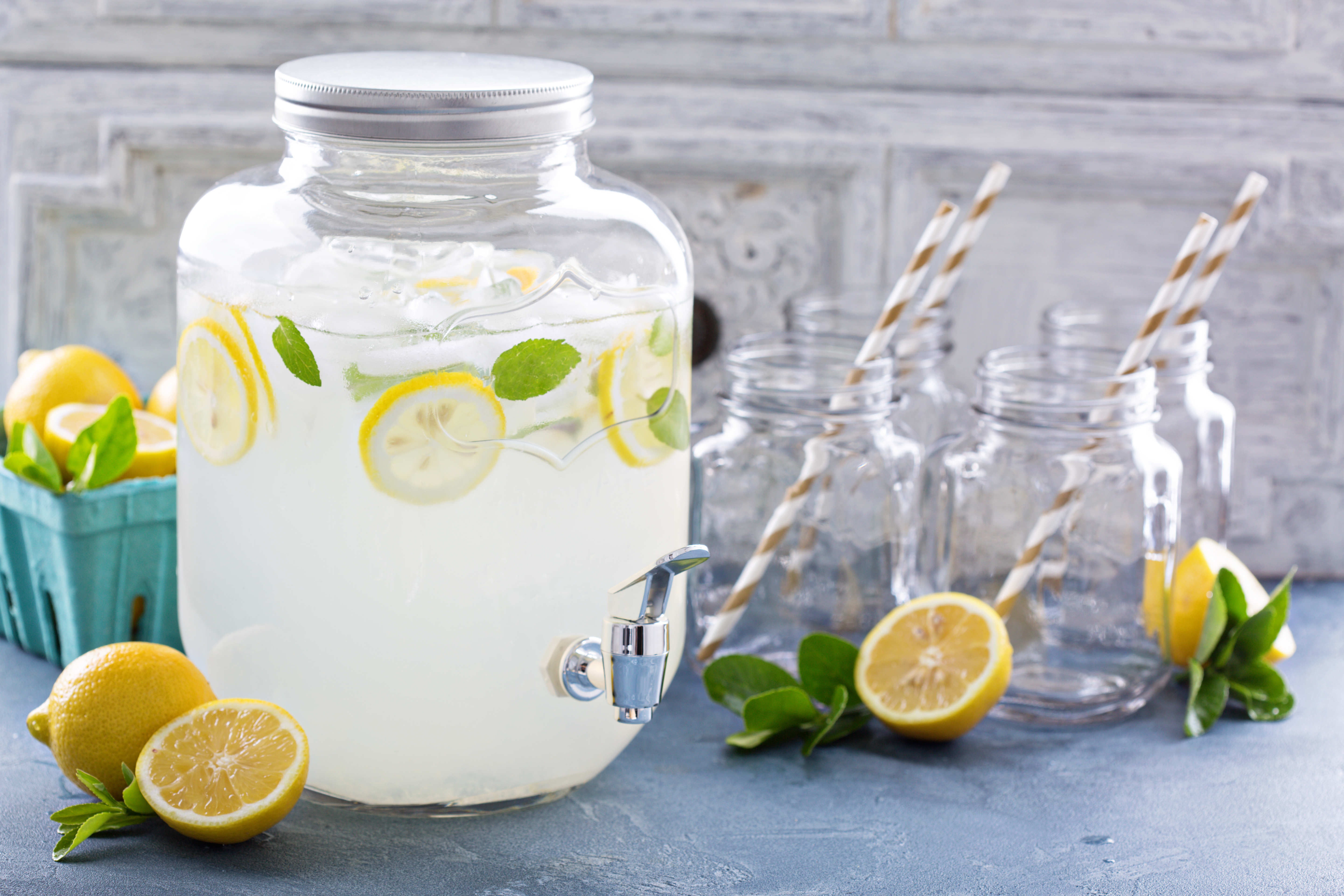 A big glass drink dispenser filled with lemonade and sliced lemons, surrounded by drinking glasses and sliced lemons.