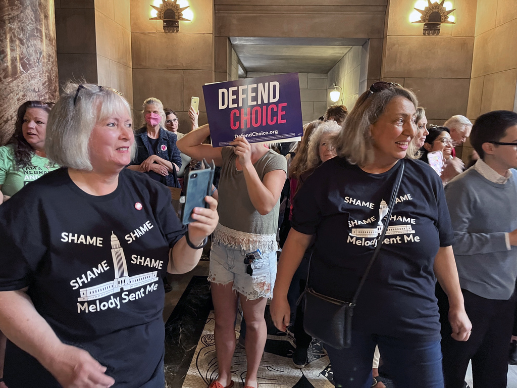 A crowd of people stands in a room. One person holds a protest sign that says “Defend Choice.” Two women, Pat and Neal, stand at the forefront, wearing matching black T-shirts printed with the words “Shame, Shame, Shame, Shame: Melody Sent Me”
