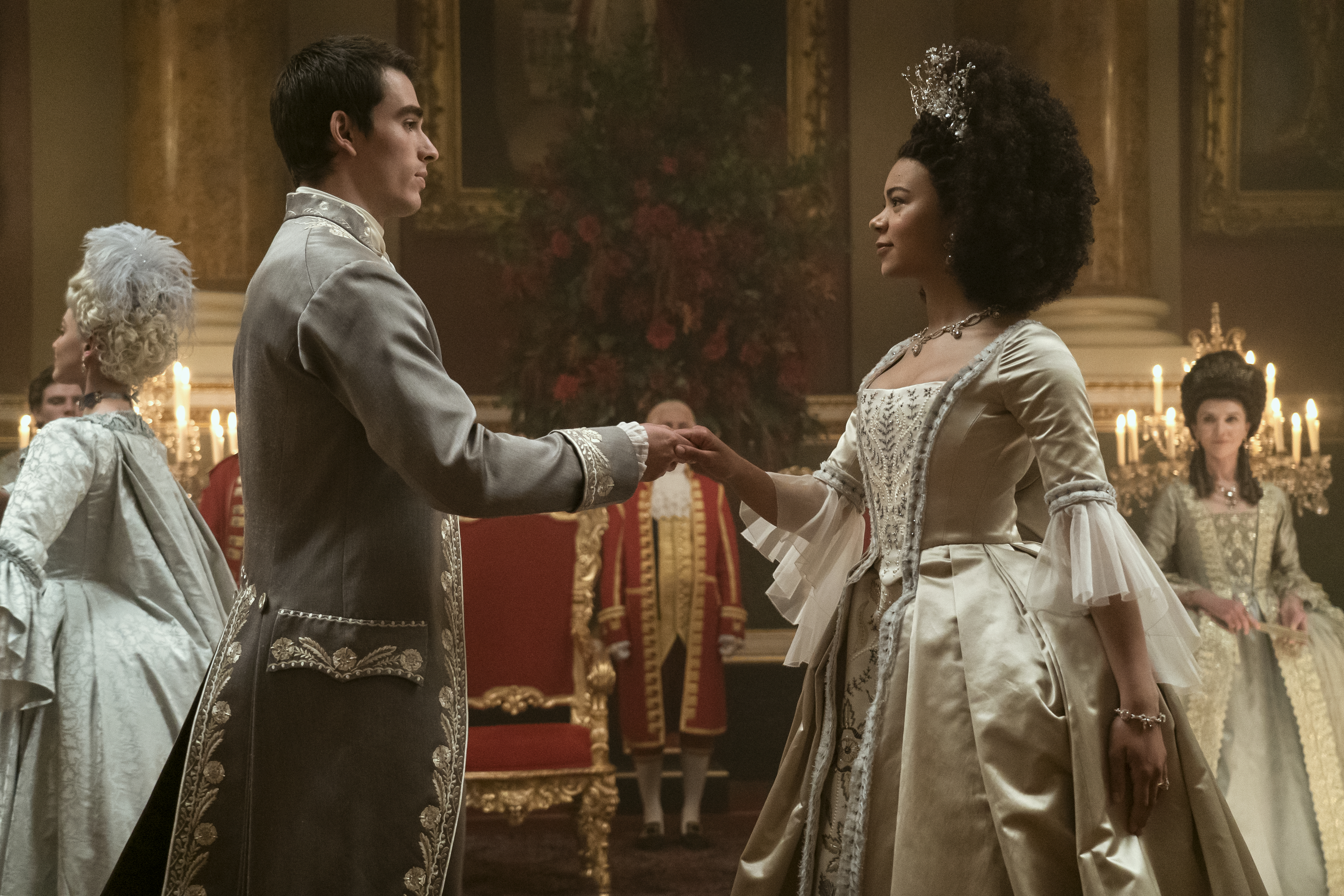 King George, a tall dark-haired young man, holds a hand out to Queen Charlotte, a young woman with an impressive afro