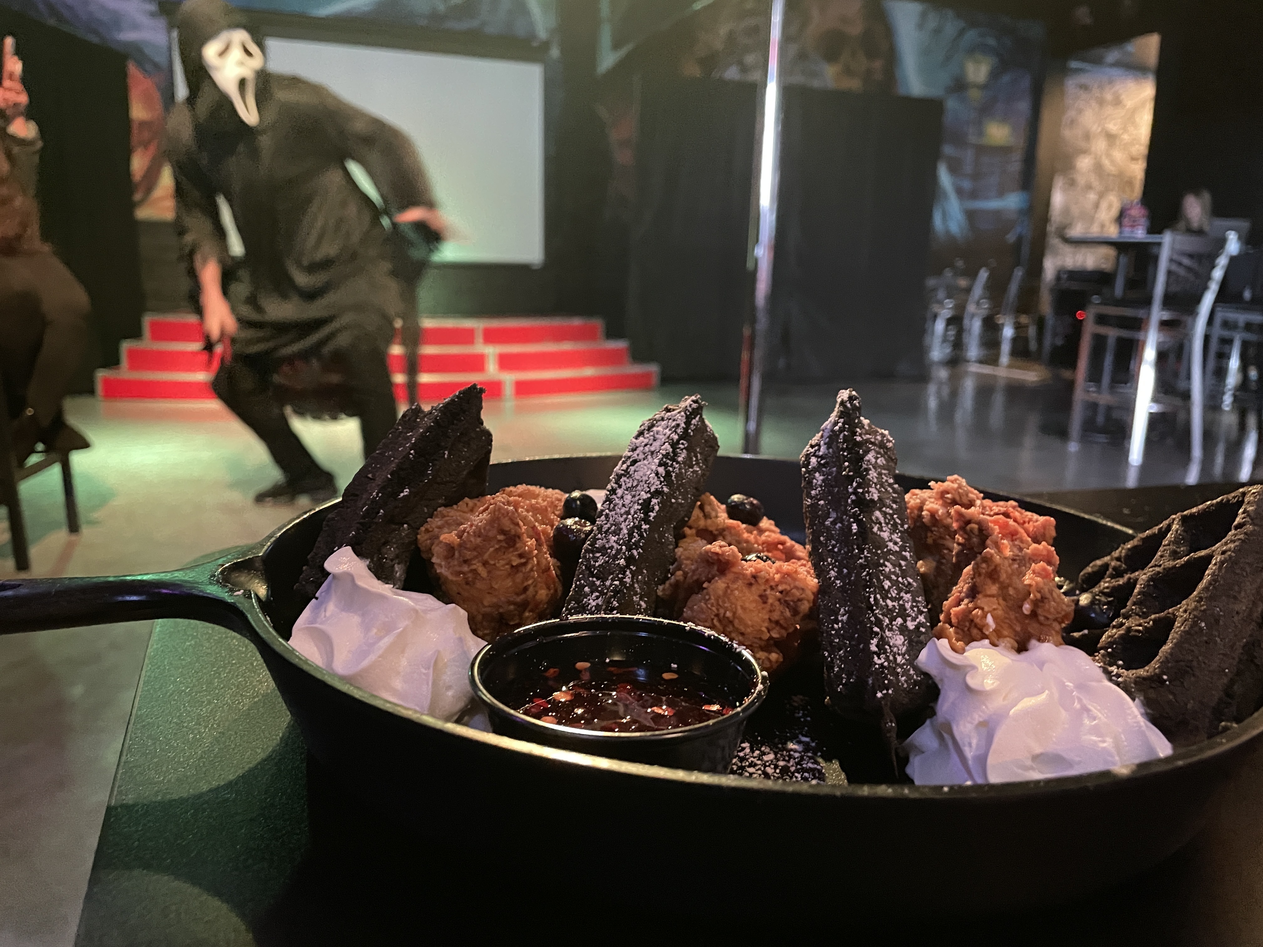 A hooded figure performs behind a plate of chicken and waffles.