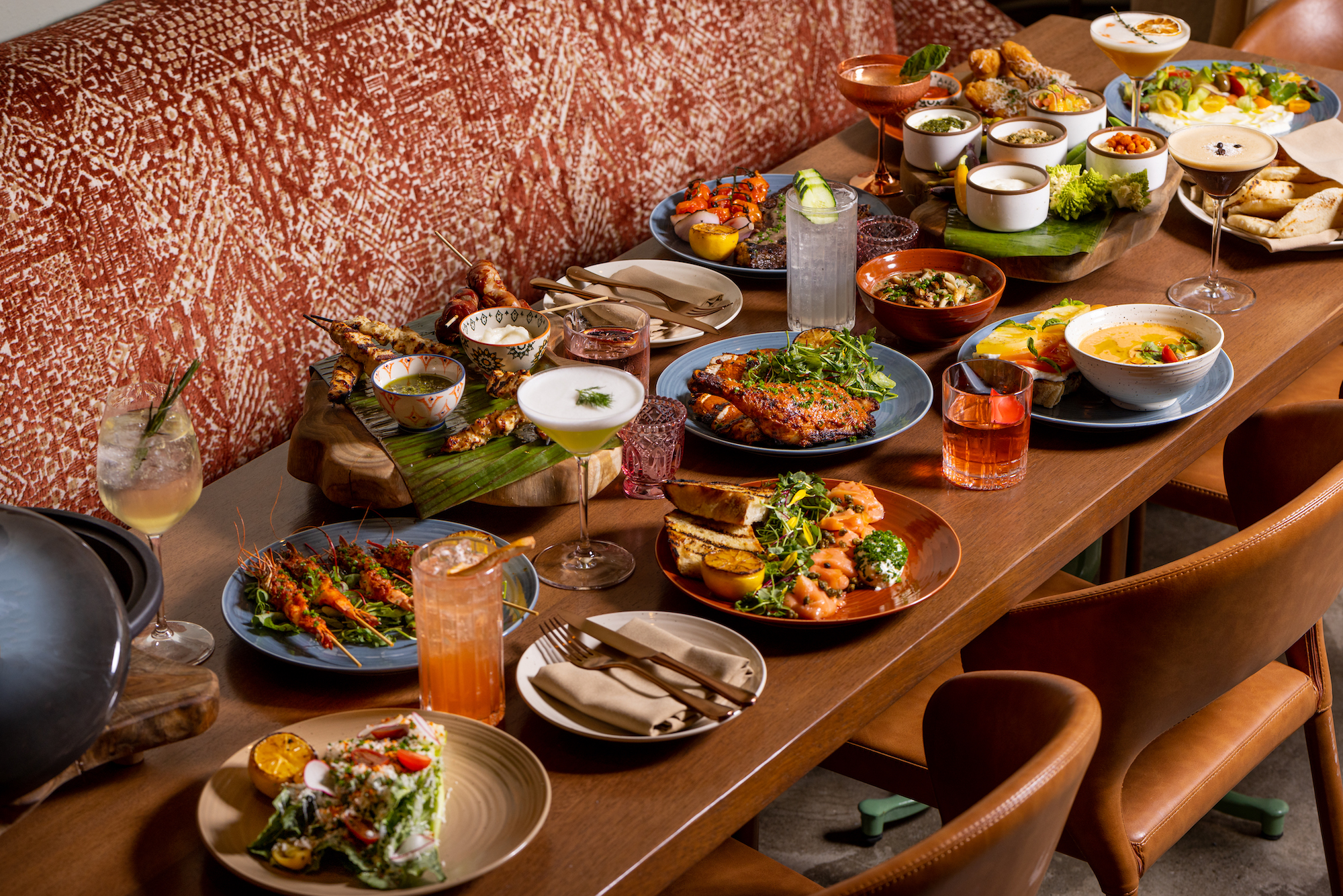 A side angle of a long wooden table filled with meats and vegetables and cocktails at a new restaurant at daytime.
