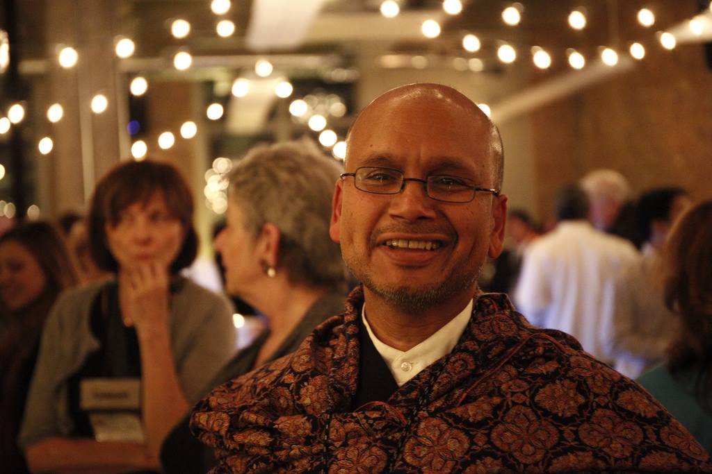 Raghavan Iyer, wearing glasses with a shawl draped around his shoulders, smiles. In there background there are people talking and strings of lights.