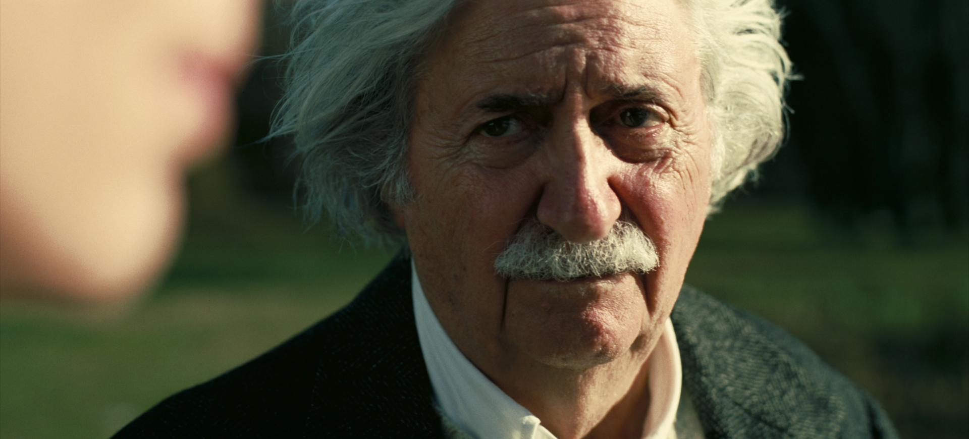 A close up of Albert Einstein, as seen in Christopher Nolan’s Oppenheimer. He is an older man with an impressive white mustache and fluffy white hair.